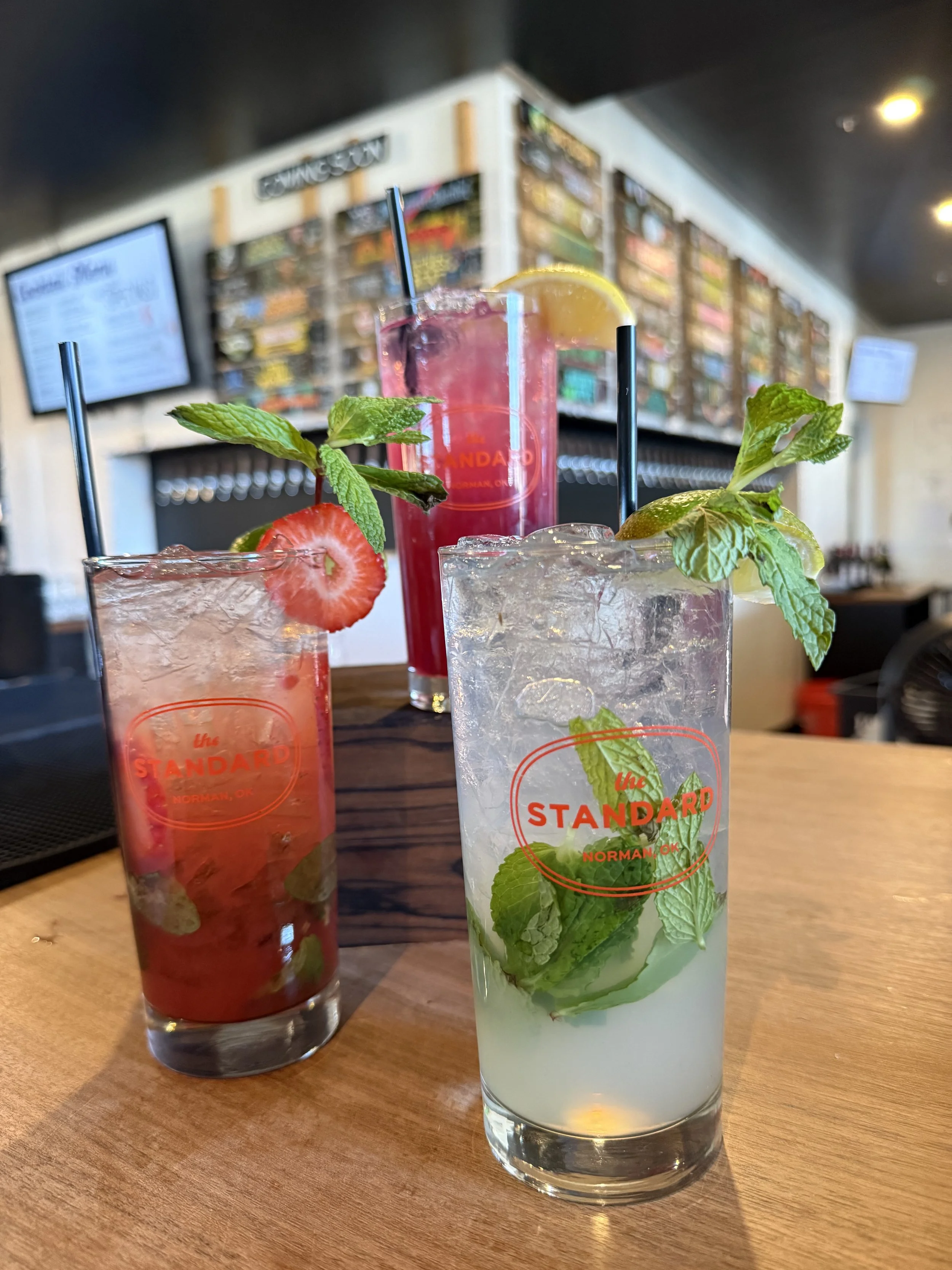 Three colorful cocktails with mint and fruit garnishes on a wooden bar counter at a restaurant.