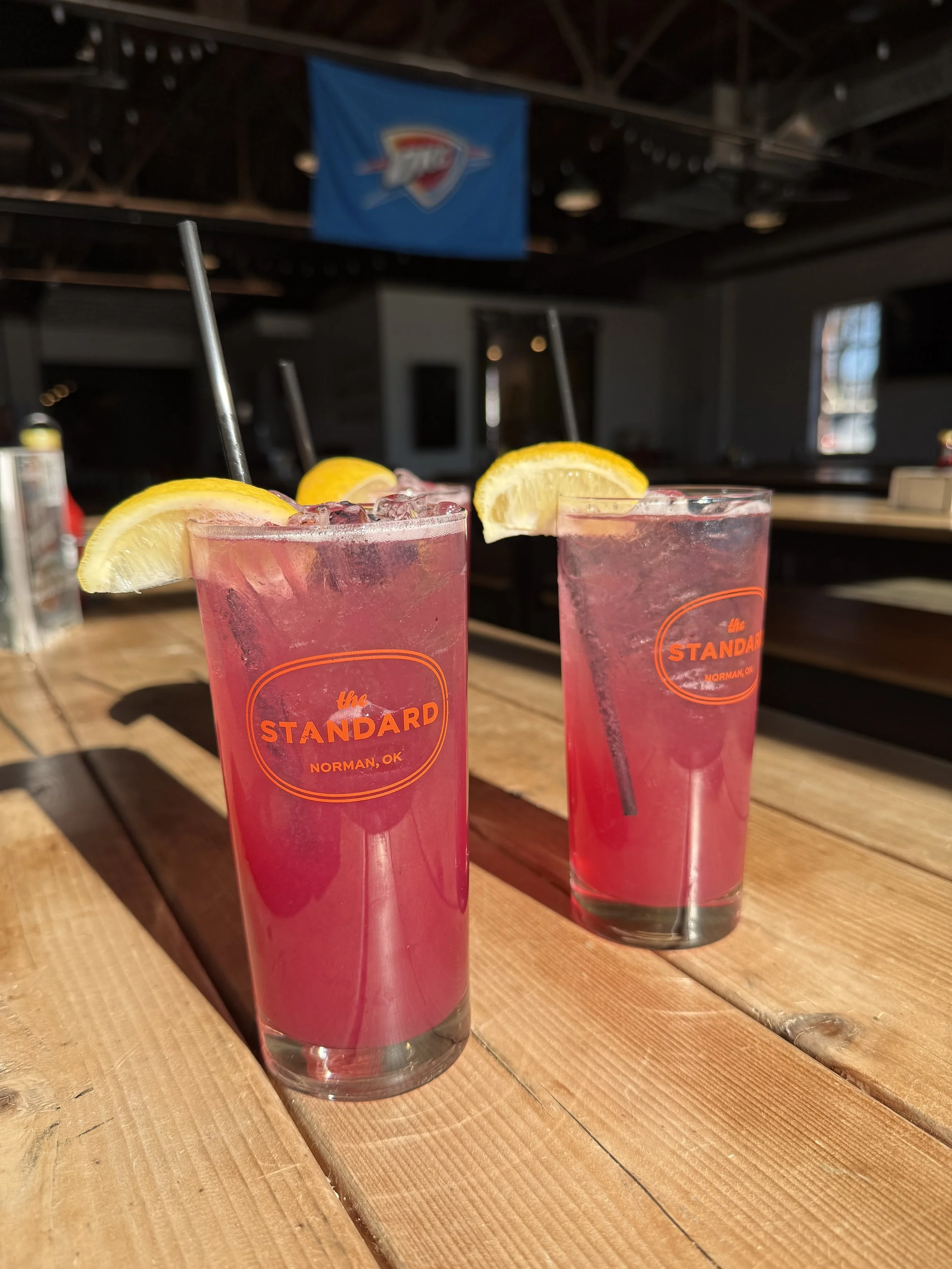 Two tall glasses of pink lemonade with lemon wedges, ice, and black straws on a wooden bar top at The Standard in Norman, Oklahoma.