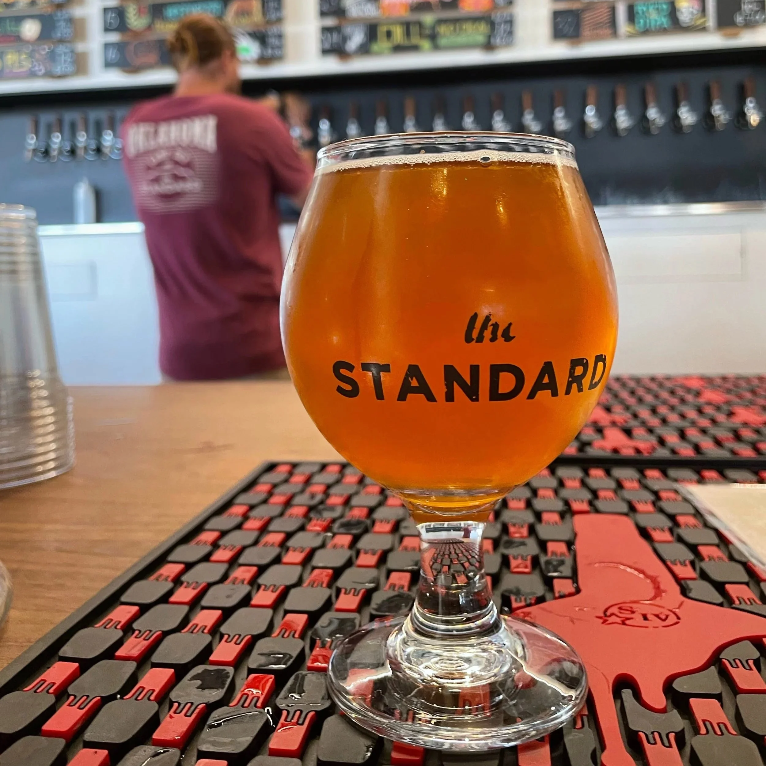 A glass of amber-colored beer on a bar counter with a black and red bar mat, with a person working behind the bar in the background.