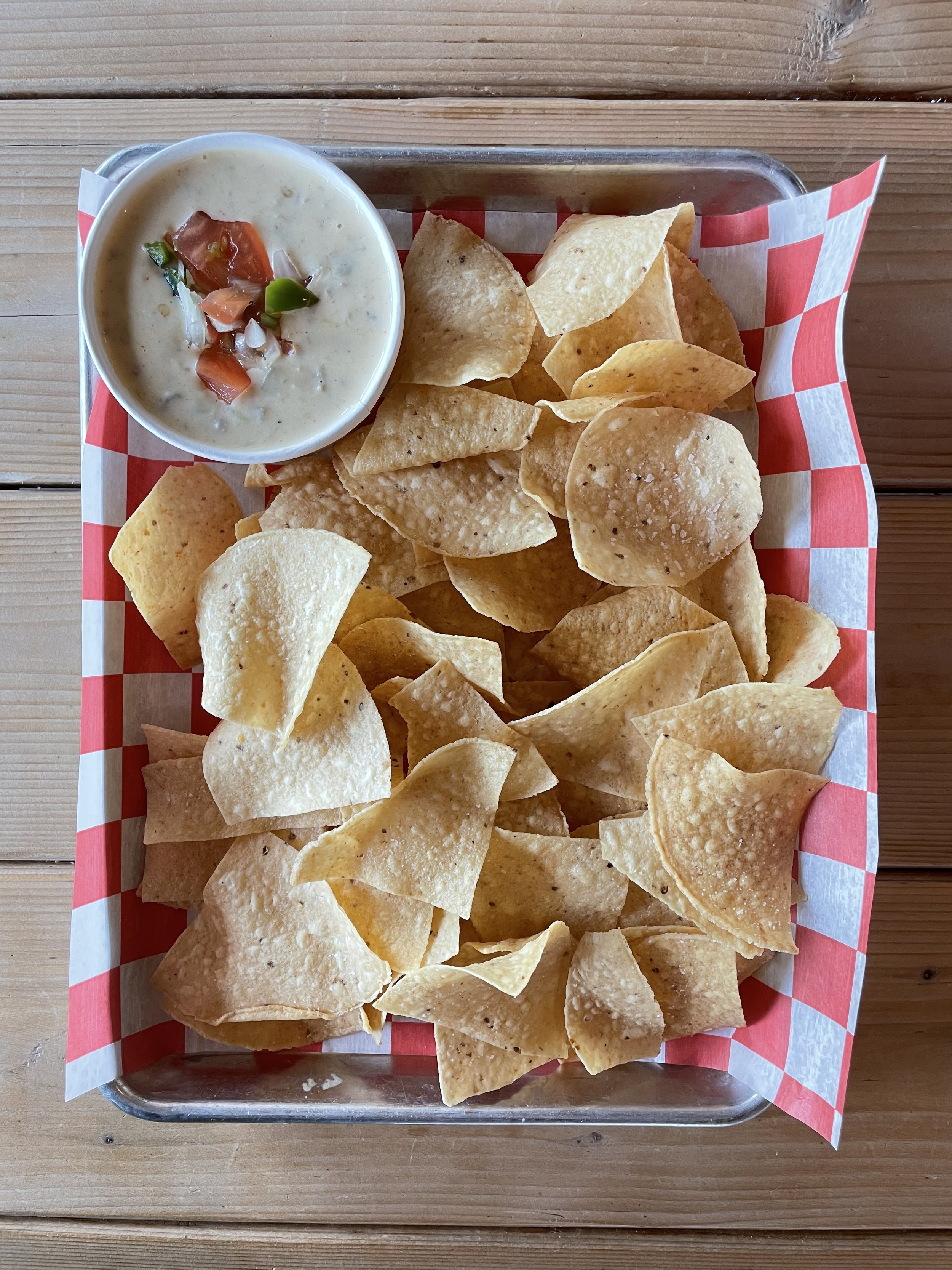 A tray of tortilla chips with a small cup of creamy dip topped with chopped tomatoes and green peppers, on a wooden table.