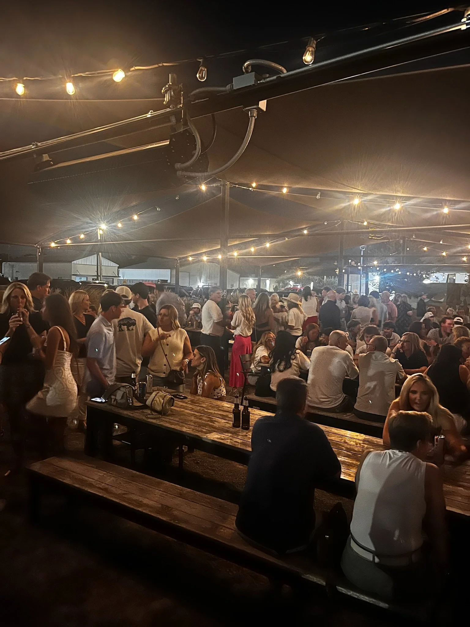 A crowded indoor party or event with people socializing, sitting at wooden tables, and standing under string lights.