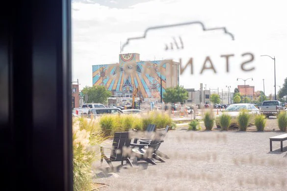 View through a window showing a parking lot, benches, greenery, and a mural on a building in the distance.