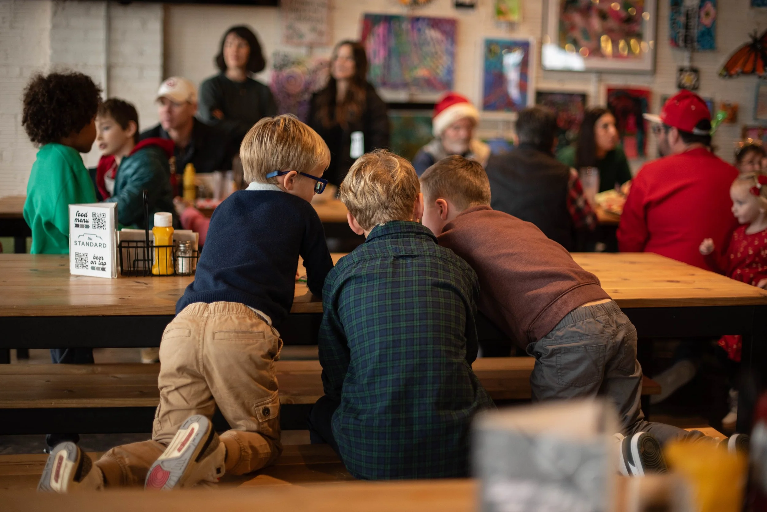 A group of children crouched down and talking at a wooden table in a lively, decorated restaurant during a holiday celebration.