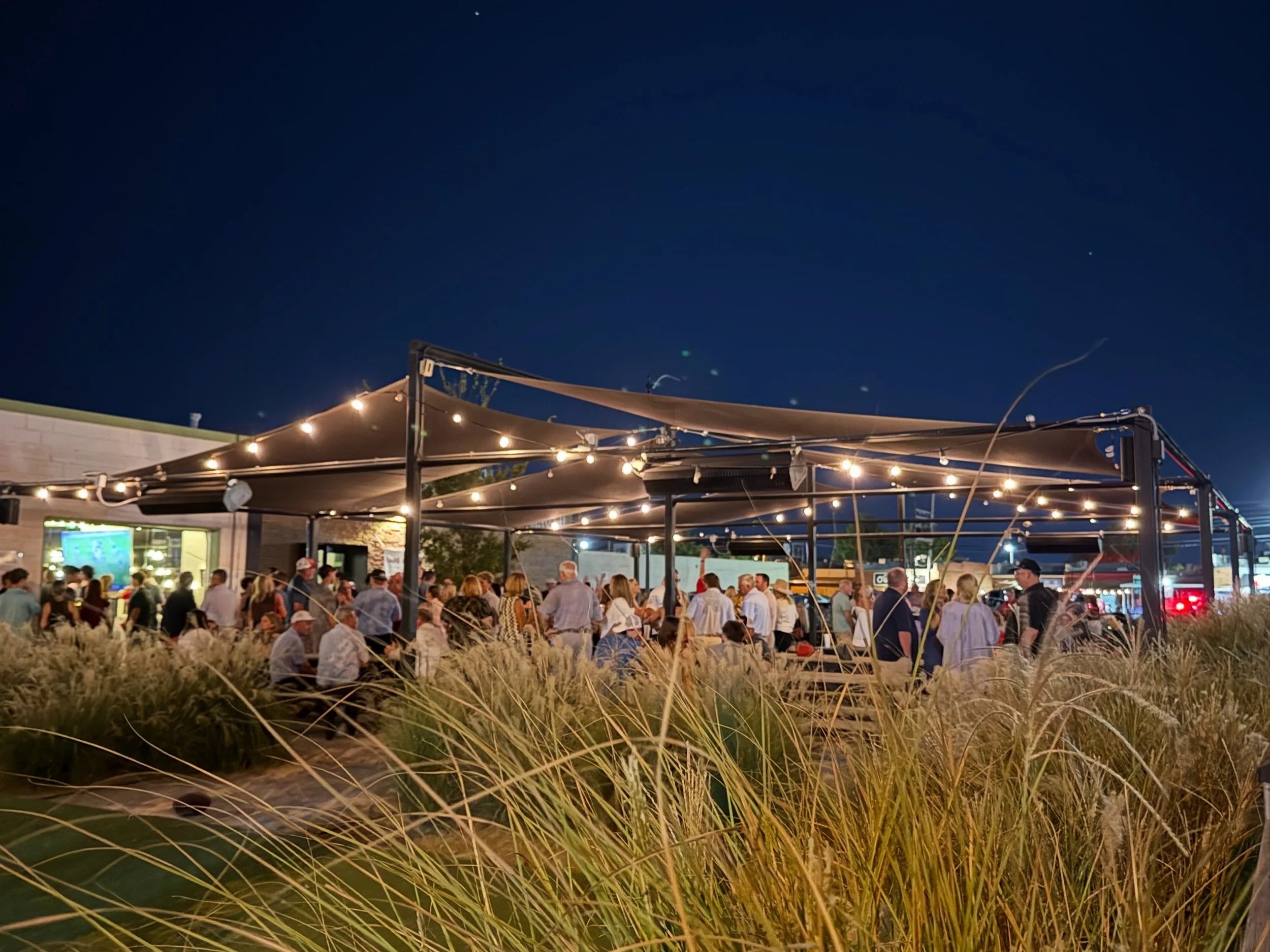 An outdoor gathering at night with people under a canopy of string lights in a modern outdoor space with grass in the foreground.