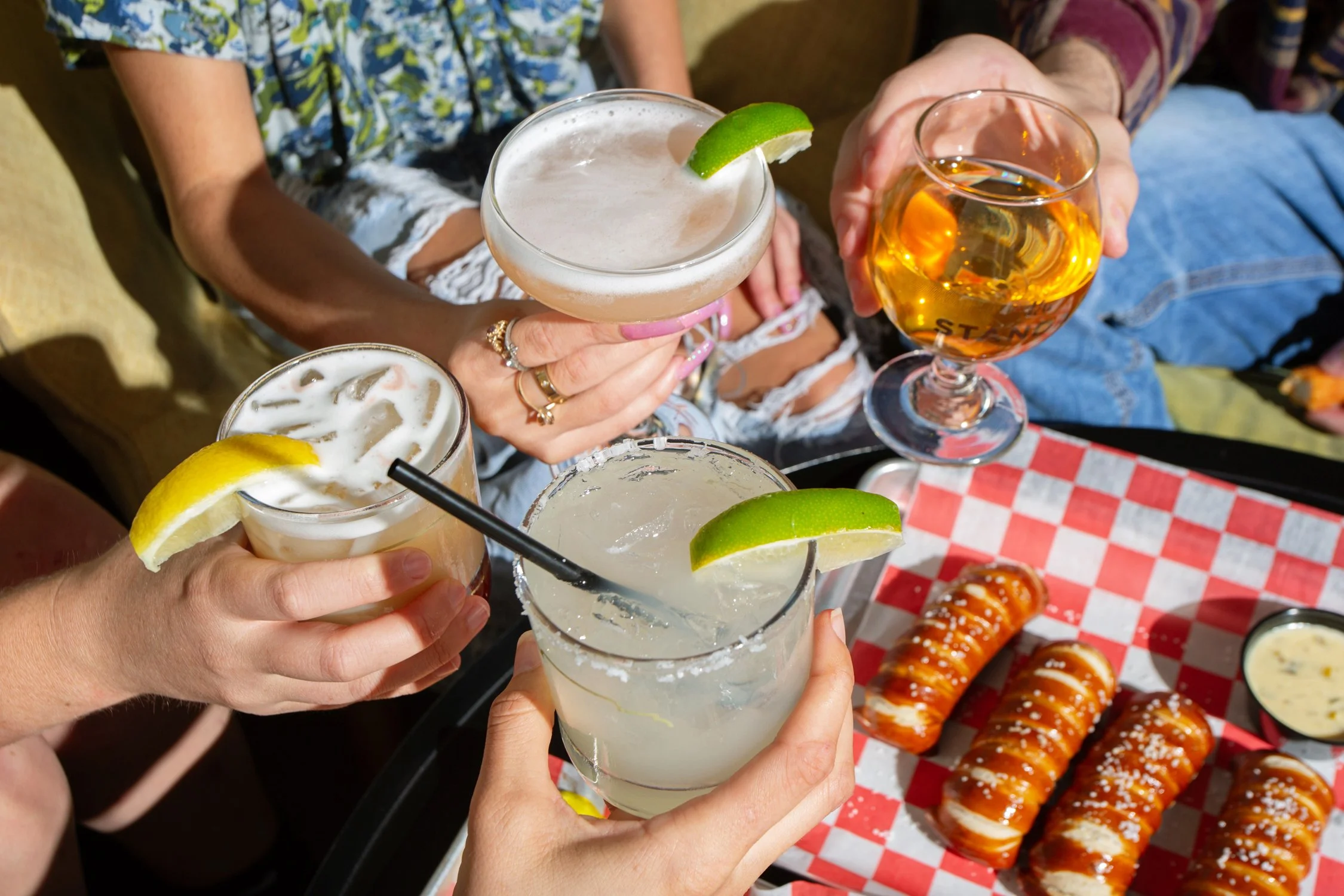 People holding various cocktails and beer glasses with lime and lemon garnishes at a gathering, with food on a checkered paper in the background.