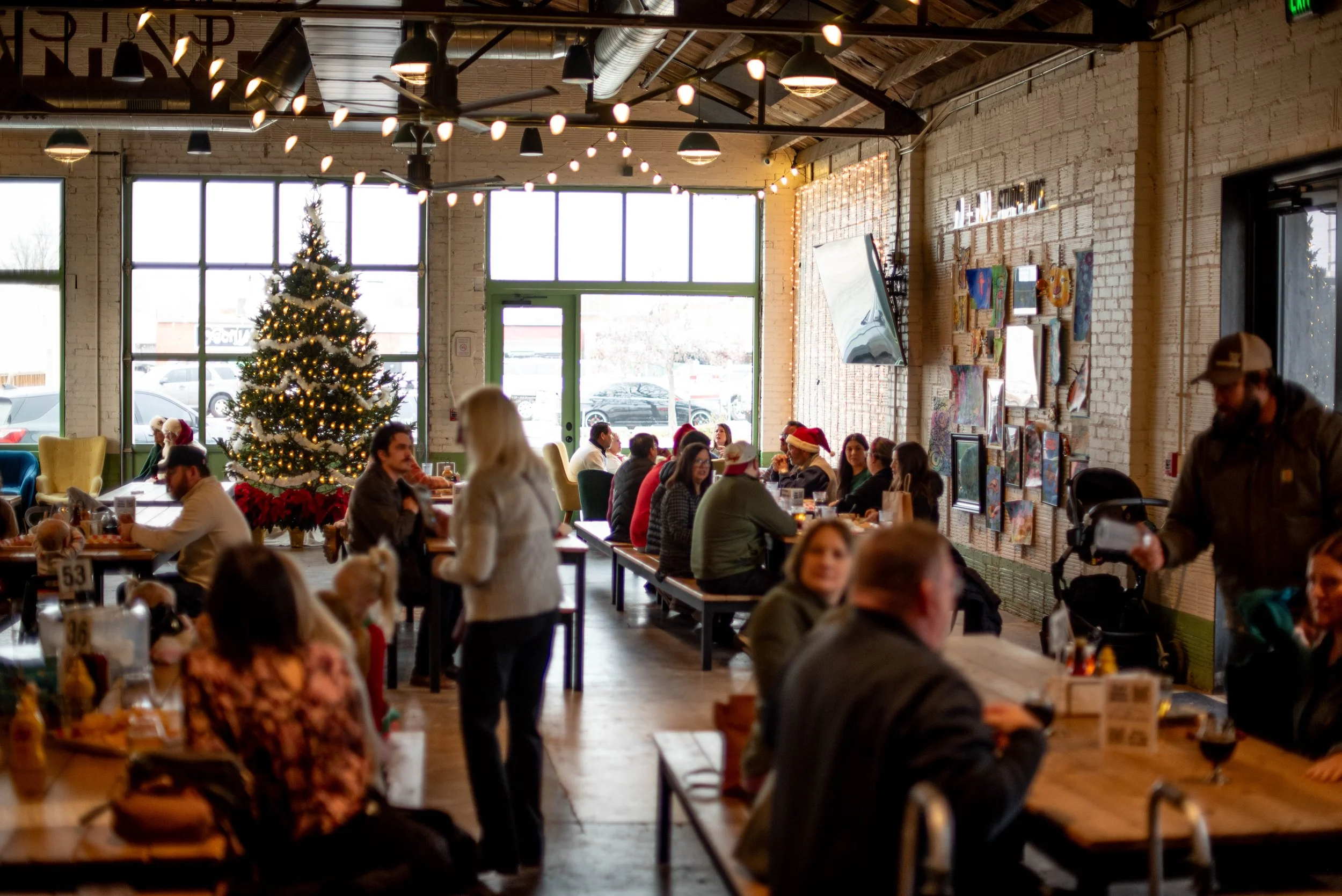 A festive indoor gathering in a restaurant decorated for Christmas with a large decorated Christmas tree, holiday lights, and people socializing at tables, some wearing Santa hats.