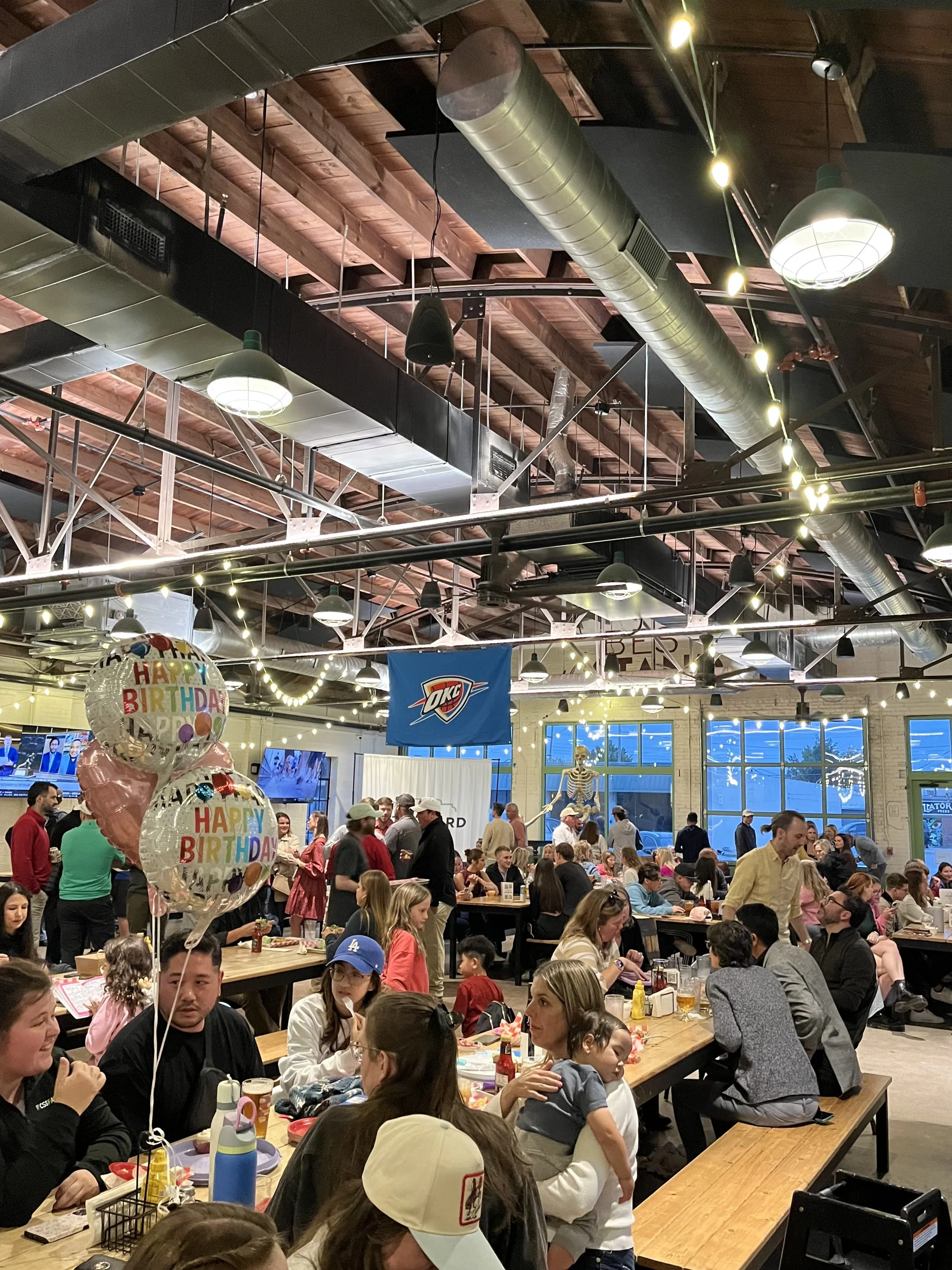 A lively indoor birthday celebration with many guests seated at tables, balloons, and a skeleton decoration in the background.
