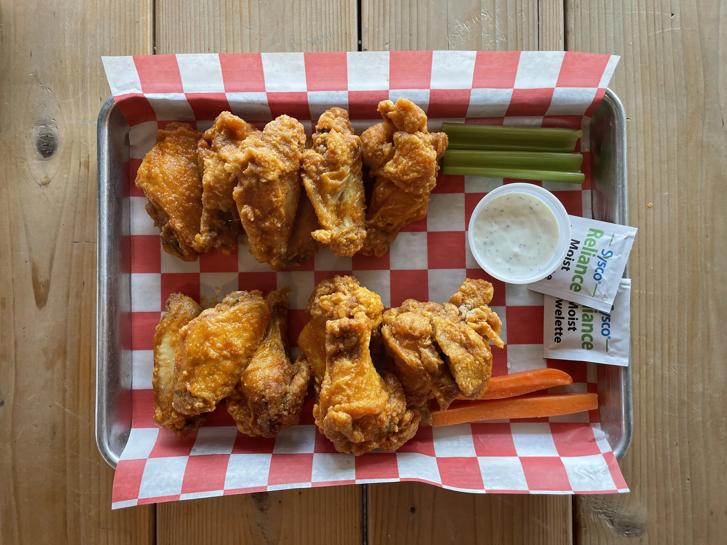 A metal tray with fried chicken wings, celery sticks, carrot sticks, a small cup of ranch dressing, and packets of saltine crackers on a wooden table.