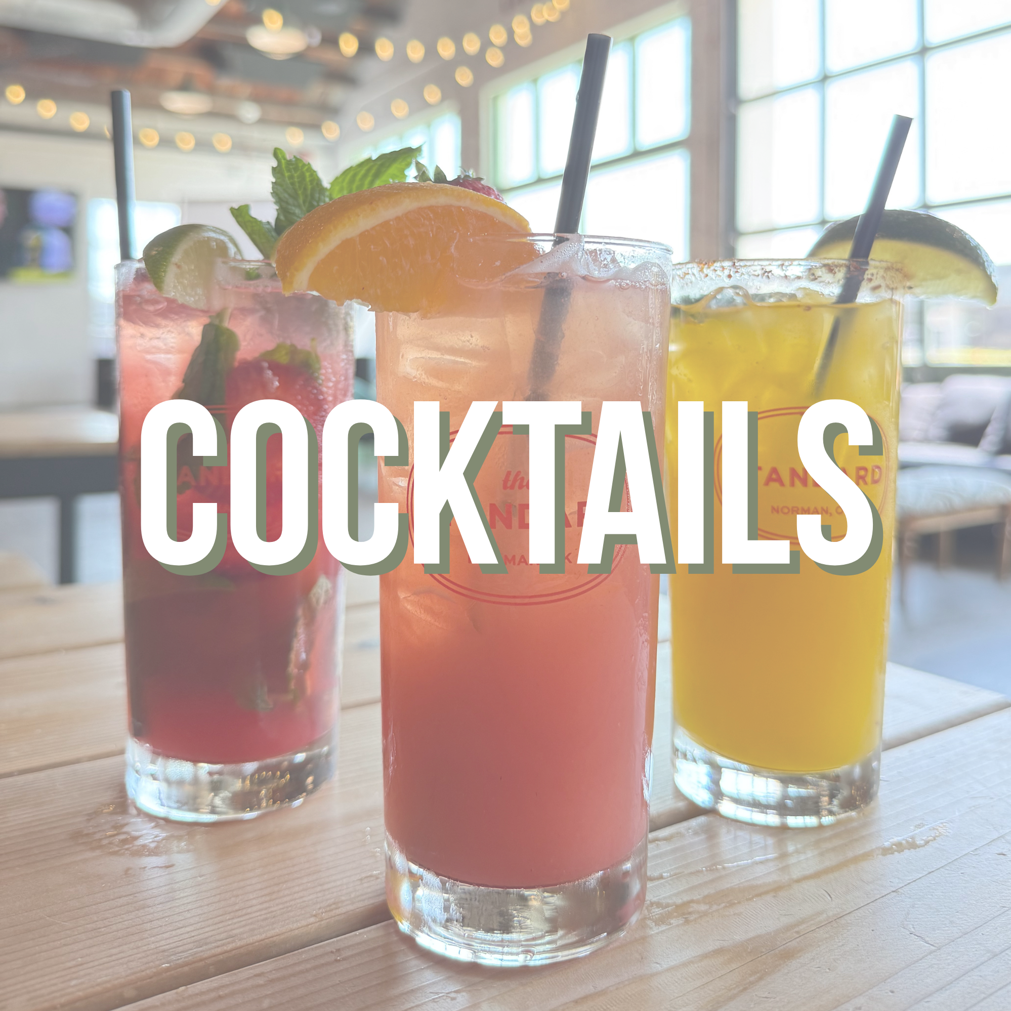 Three colorful cocktails in tall glasses with straws and garnishes on a wooden table in a bright, modern restaurant.