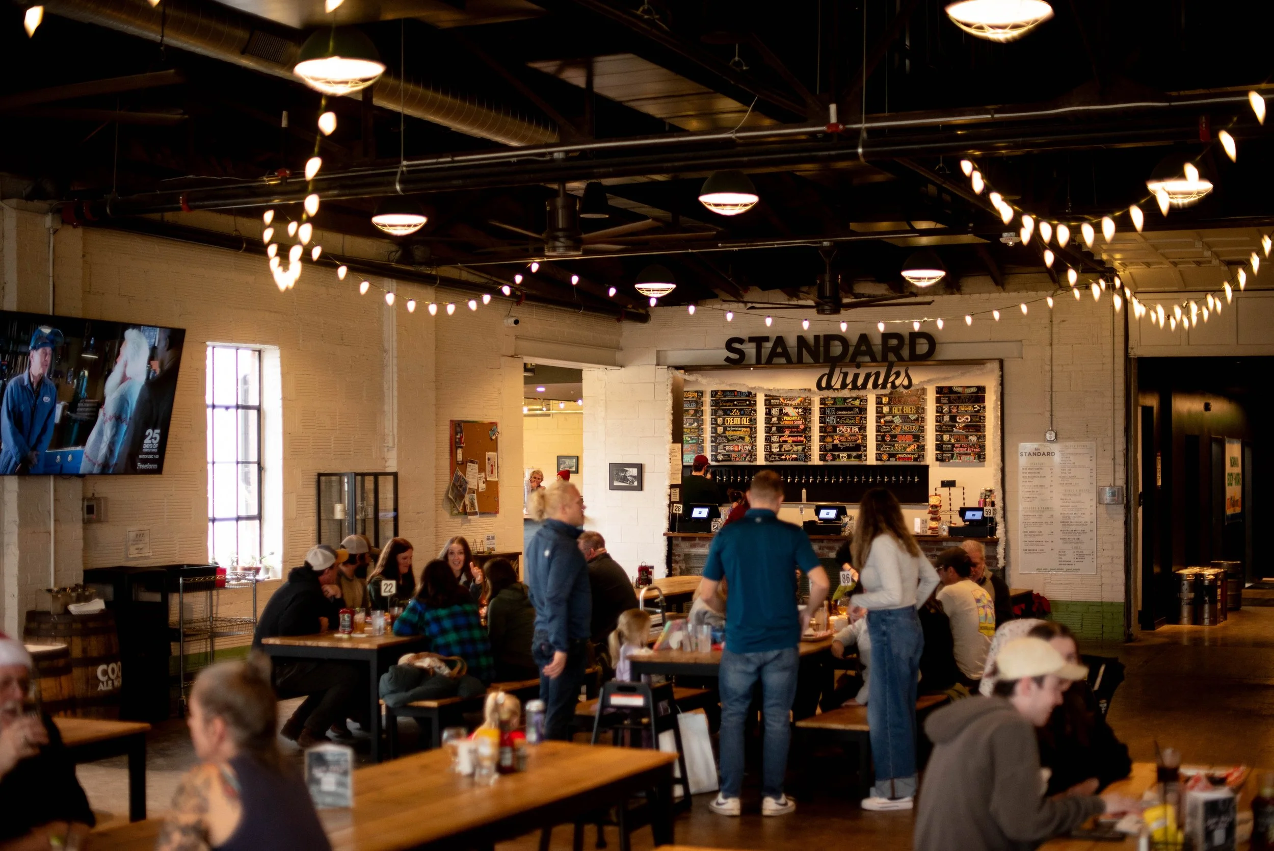 People dining inside a brewery or bar with string lights and a menu board labeled 'STANDARD drinks' in the background.