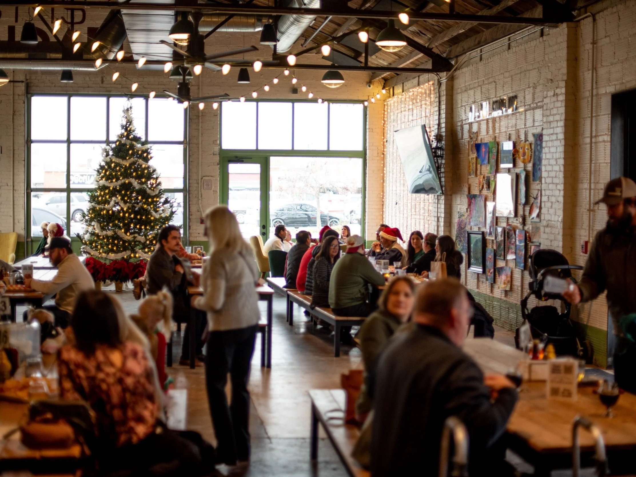 A decorated indoor space with a Christmas tree, holiday decorations, and people dining and socializing, some wearing Santa hats.