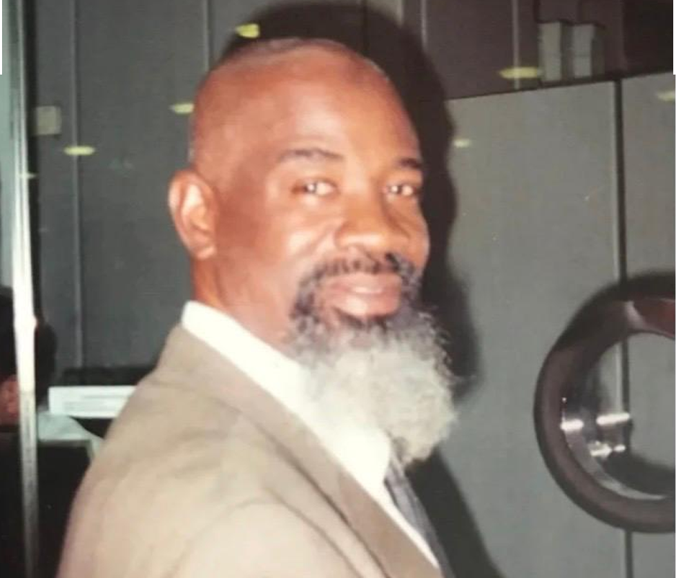 A middle-aged man with a beard and mustache, wearing a suit and tie, standing indoors near a mirror or window, looking at the camera.