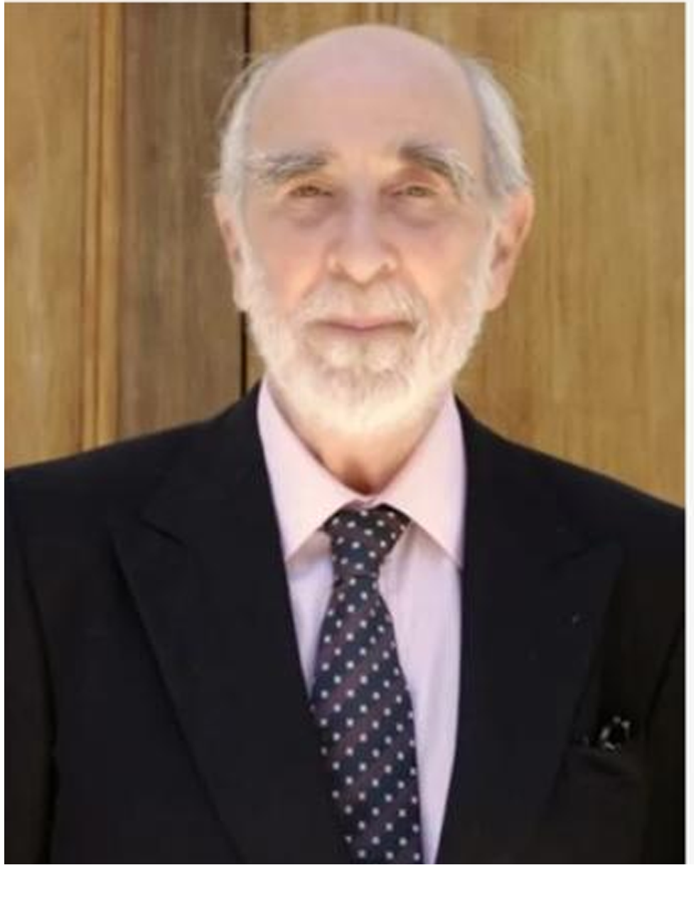 An older man with a white beard and bald head, dressed in a black suit, light pink shirt, and polka dot tie, standing in front of a wooden panel background.