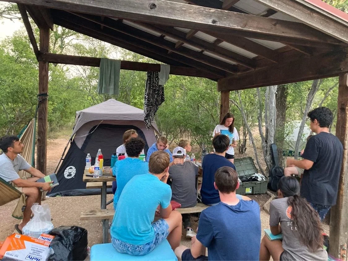Group of young people gathered around a table with a tent in a wooded outdoor setting, attending a presentation or discussion under a wooden shelter.