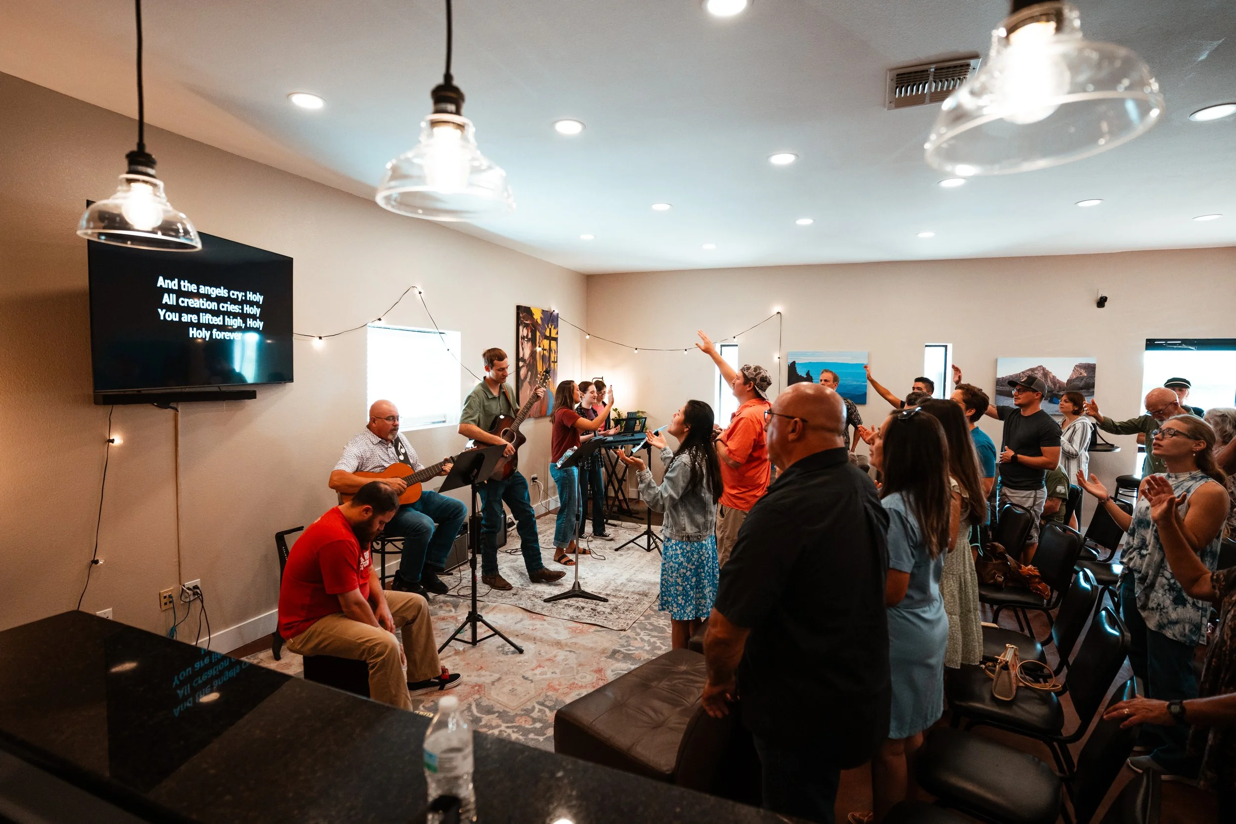 People attending a worship and communion event in a cozy room with a presenter at the front, a large screen displaying discussion questions, musical instruments, and an illuminated 'Open' sign on the wall.