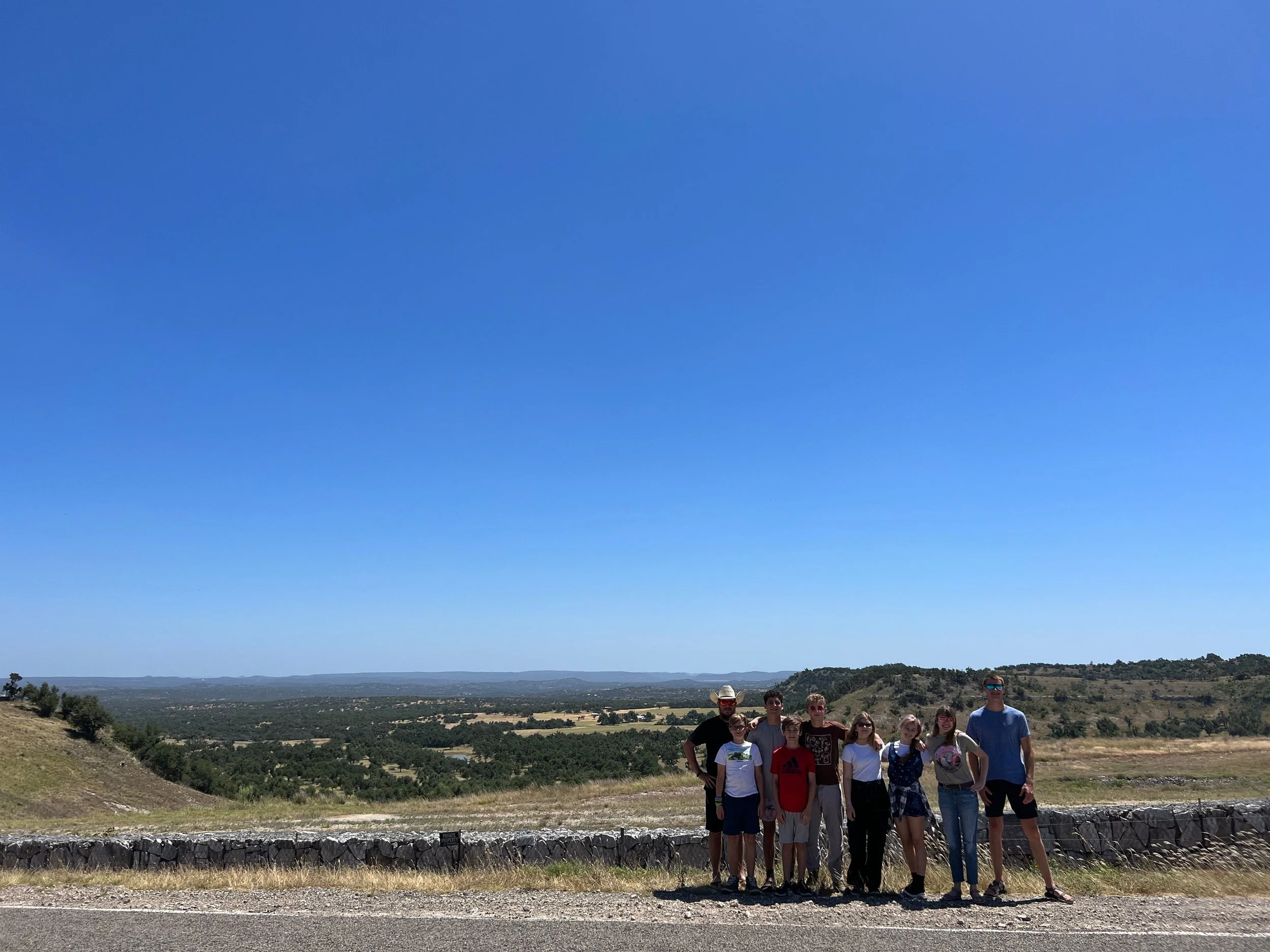 A group of nine people standing outdoors on a sunny day in front of a stone barrier with a scenic landscape of rolling hills, trees, and a clear blue sky in the background.