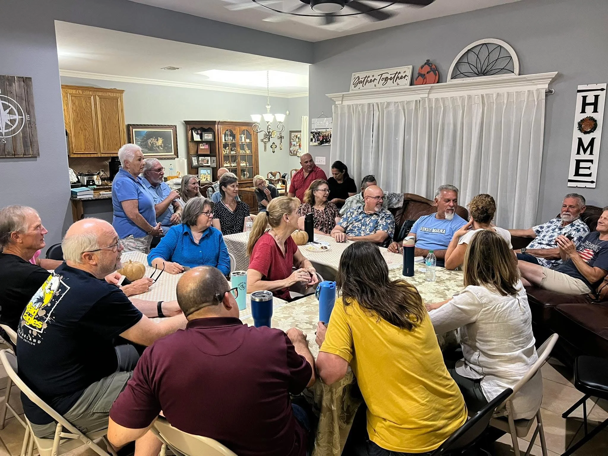 A group of people gathered around a dining table, smiling, with a hanging chandelier above, in a home setting.