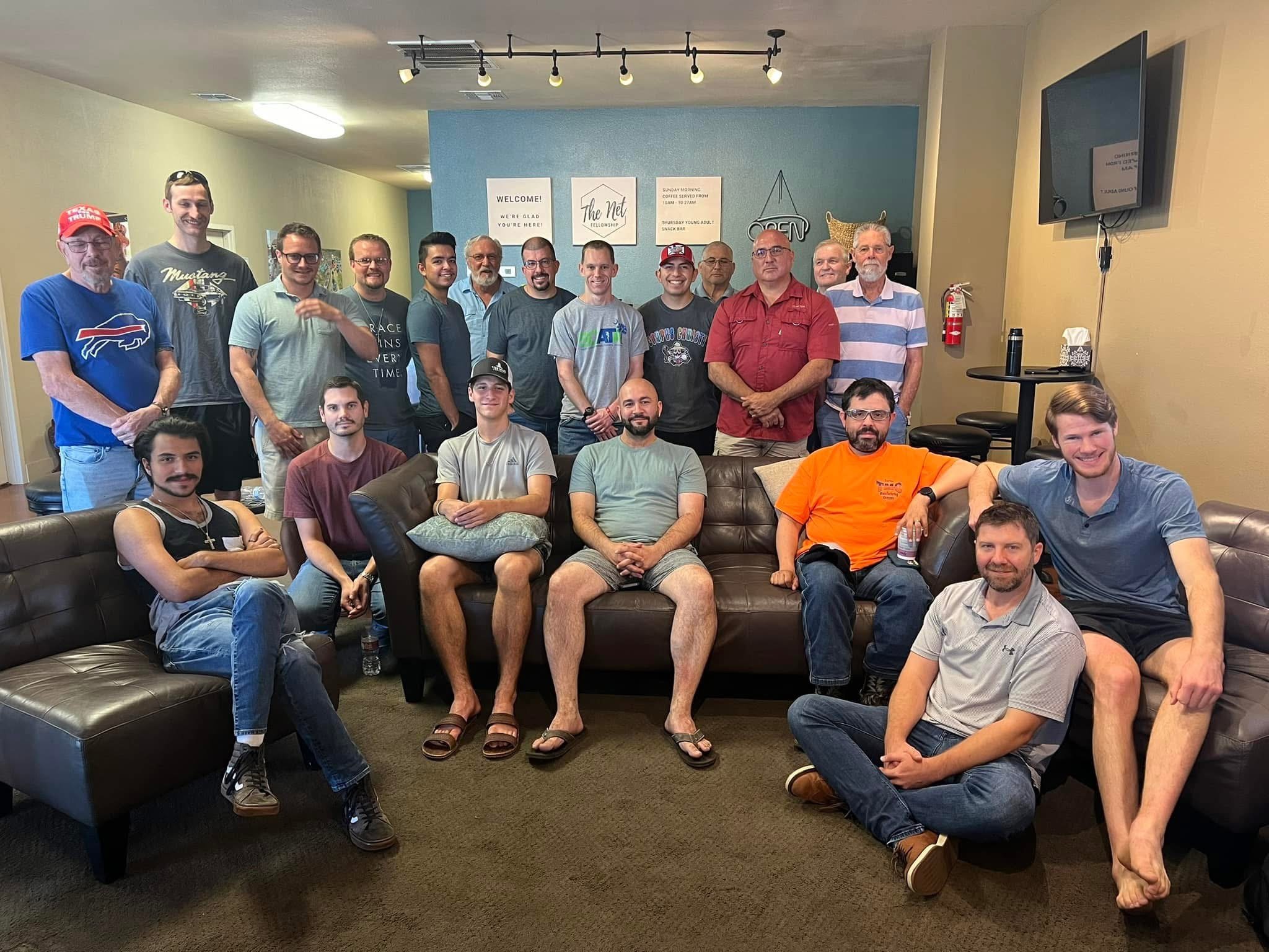 A group of men sitting around a table in a restaurant, with a woman standing and talking to them, during a gathering called 'The Net Fellowship Men's' event.