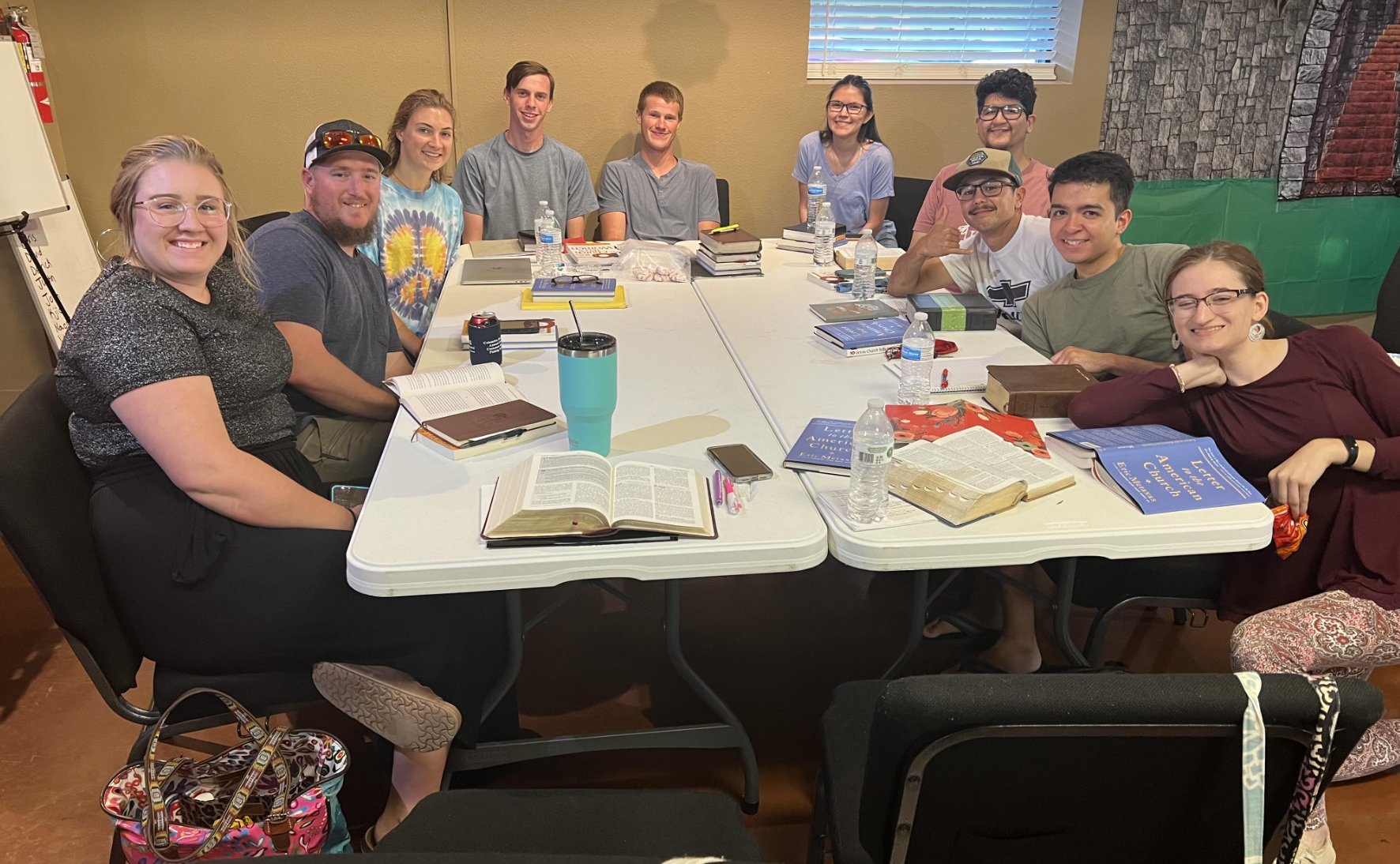 A group of fifteen young adults sitting around a large conference table with books, water bottles, and notebooks, smiling at the camera in a classroom or meeting room.