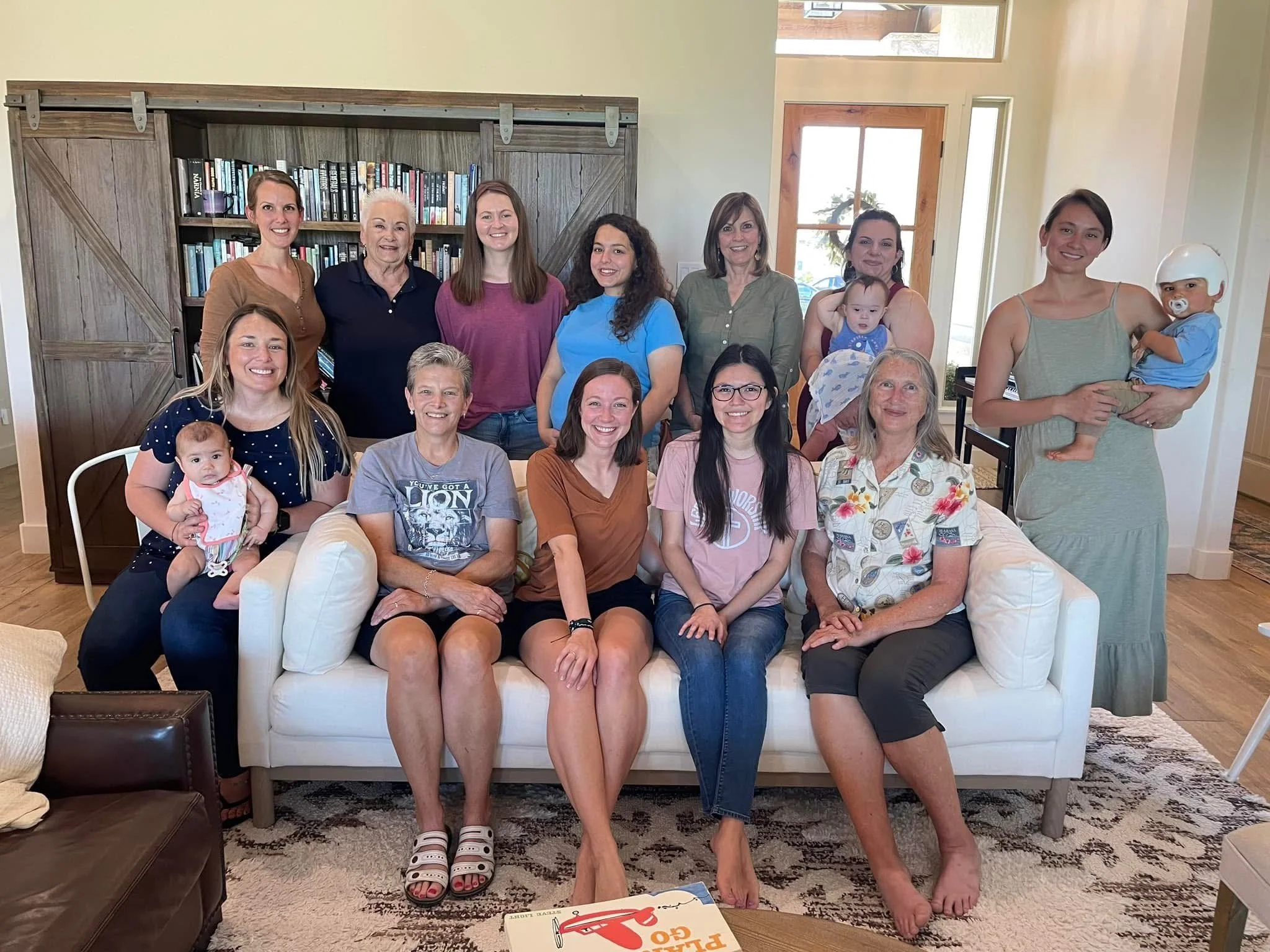 Group of women and girls gathered together in a living room, some sitting on the floor and others standing, with a large window to the right. Text overlay reads 'The Net Fellowship Women's'.