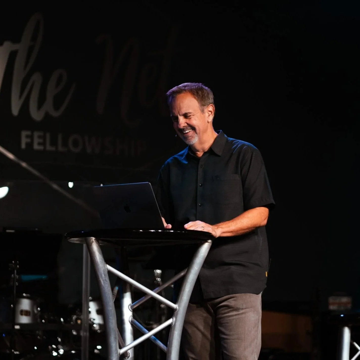 A man giving a talk or sermon at a church. Behind him is a sign that reads 'The Net Fellowship.' A water bottle and a laptop are on the table in front of him, and there are musical instruments in the background.