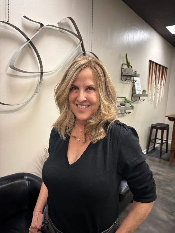 Smiling woman with blonde hair wearing a black top, standing in a modern interior with decorative wall art, shelves with plants, and a bar stool.