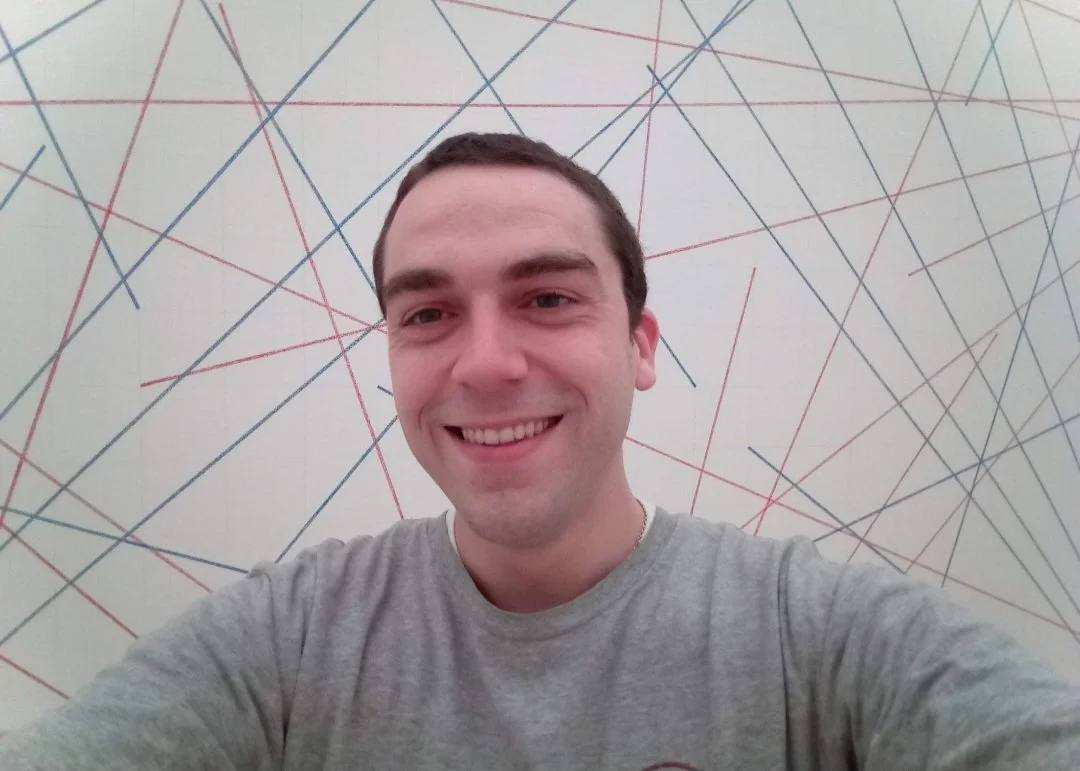 A man taking a selfie against a wall with colorful diagonal lines in blue, red, green, and yellow.