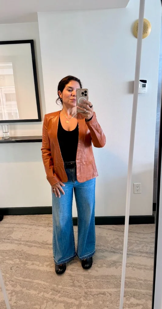 A woman taking a mirror selfie in a room with white walls and a gray carpet. She is wearing a tan leather blazer, black top, wide-leg blue jeans, black shoes, and gold jewelry.