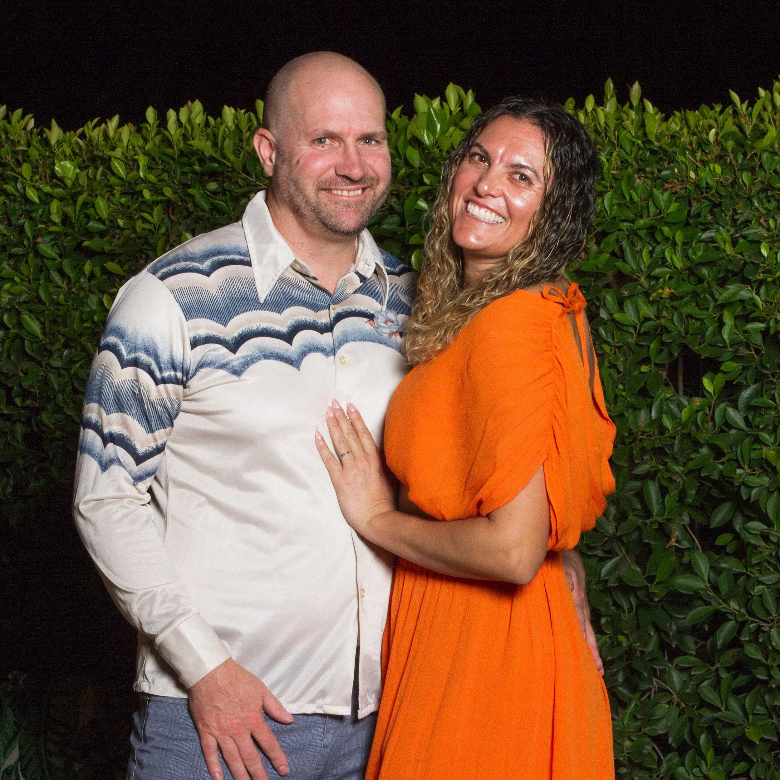 A smiling couple standing close together outdoors at night, with green bushes in the background. The man is wearing a patterned shirt, and the woman is wearing an orange dress with her hand on the man's chest.