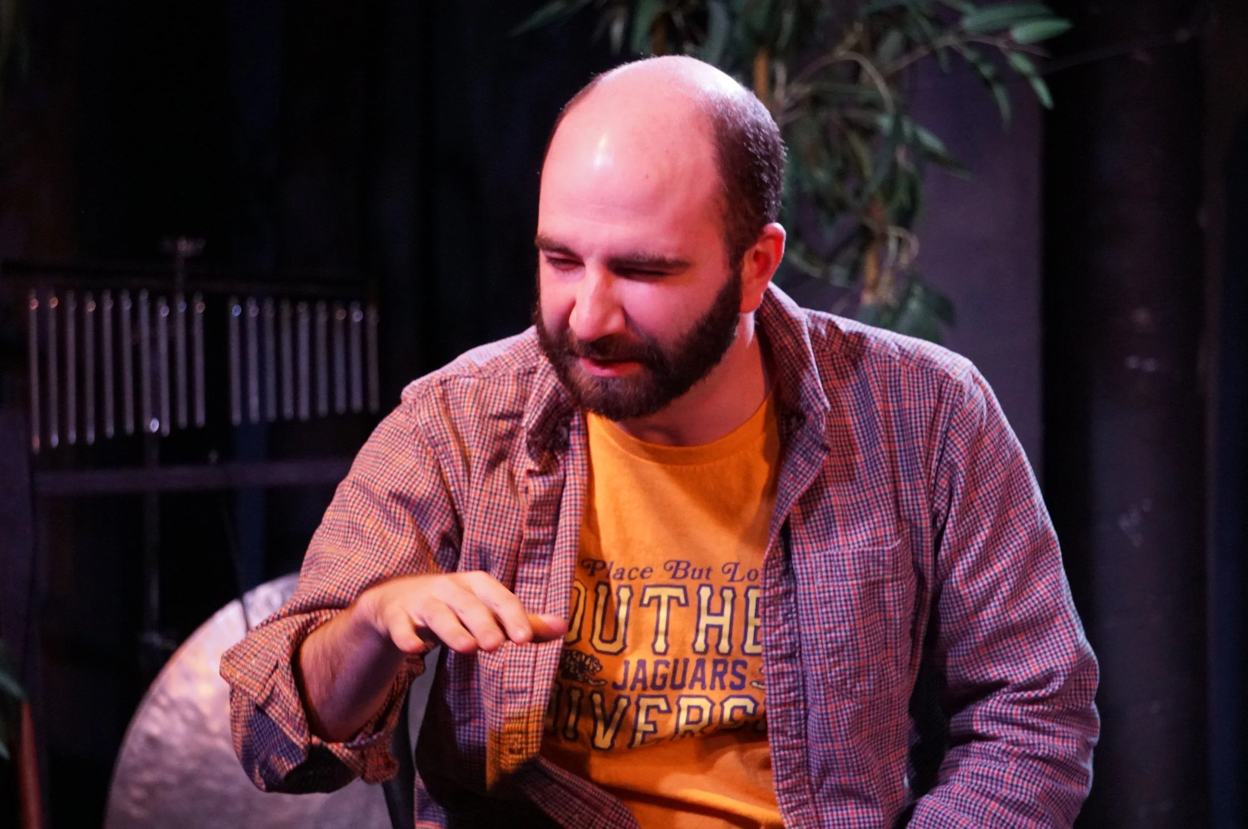 A man with a bald head and beard, wearing a yellow T-shirt and a checkered shirt, appears to be in a performance or speaking, gesturing with his hand against a dimly lit background with some plants and structures.