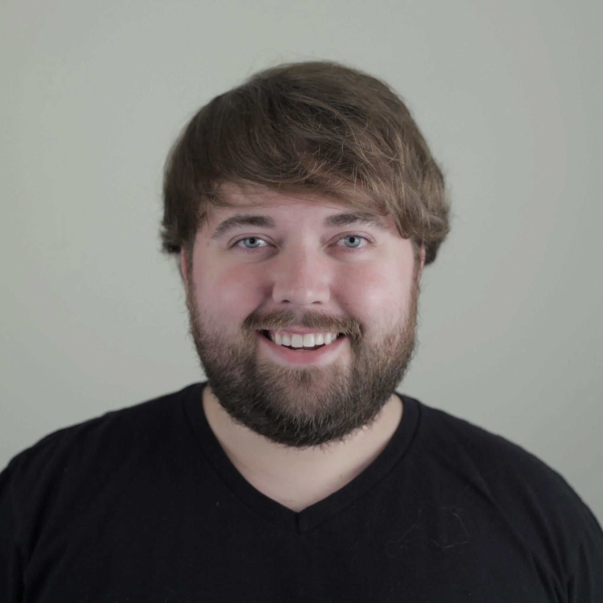 Headshot of a smiling young man with a beard and light brown hair, wearing a black shirt, against a plain light background.