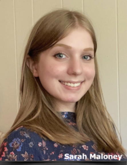 Young woman with long light brown hair and blue eyes smiling, wearing a dark floral top, standing against a light-colored wooden wall.