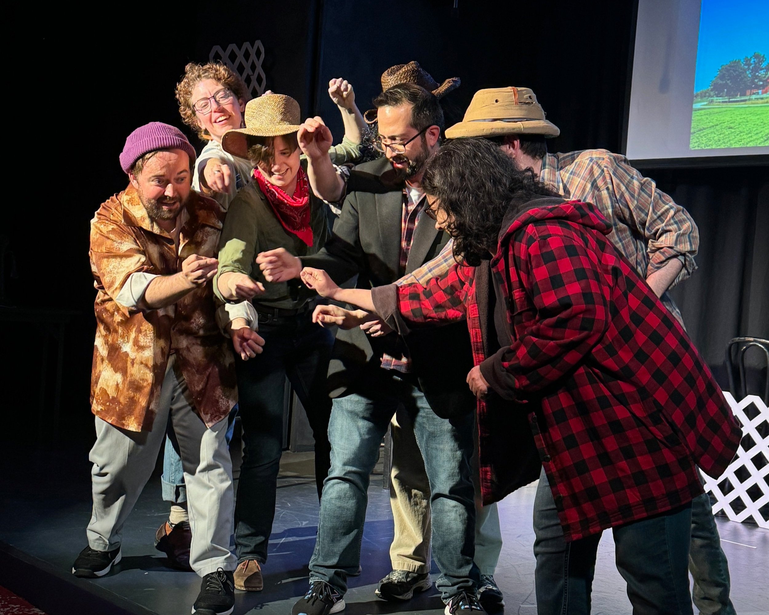 Group of actors on stage performing a play, wearing various costumes including hats and casual clothing, with some smiling and engaged in a scene.