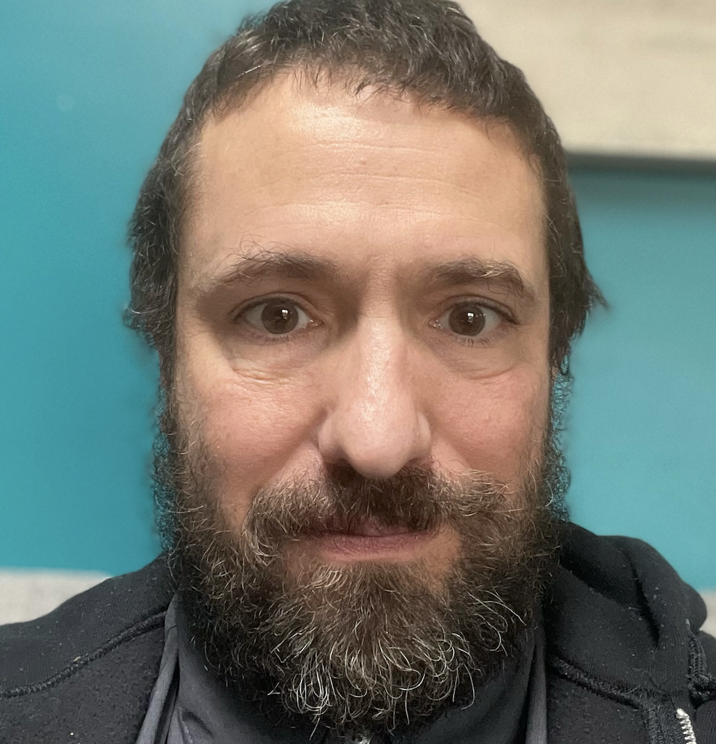 A close-up selfie of a man with brown eyes, a full beard, and short dark hair, wearing a black jacket against a blue background.