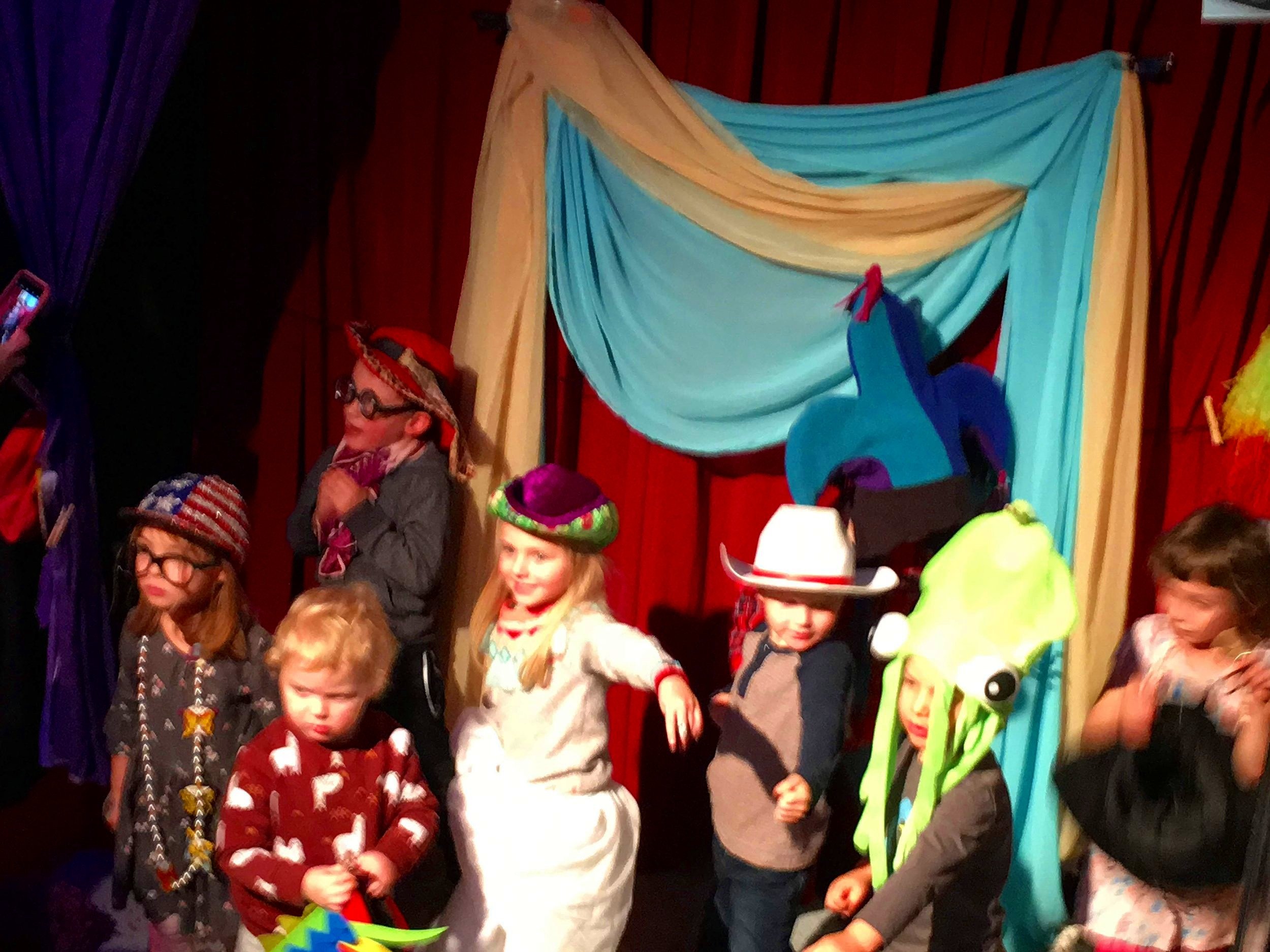 Children in costumes performing on a stage with a colorful backdrop featuring a blue and yellow curtain and a guitar silhouette.