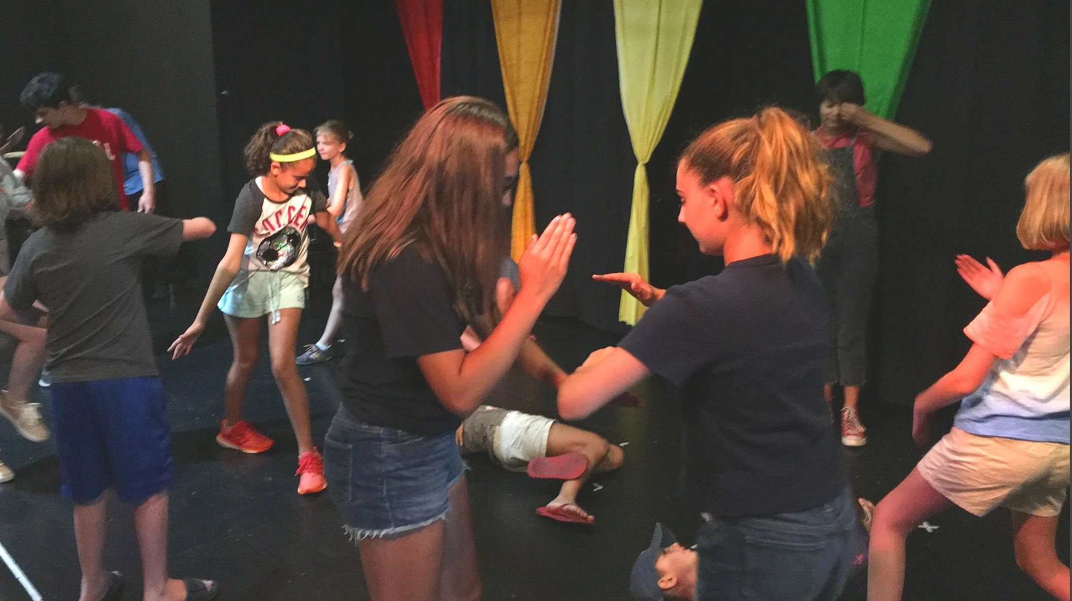 Group of children dancing and playing on a stage with black background and colorful hanging fabric.