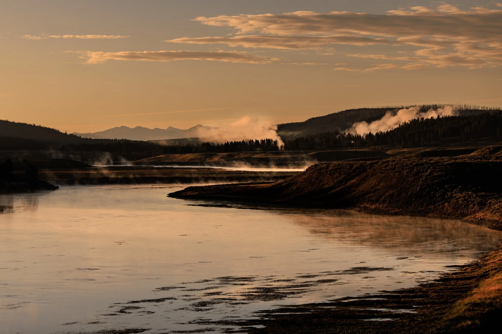 The Yellowstone River winding through Hayden Valley at sunrise, with steam rising from the water and thermal areas and distant mountains on the horizon.