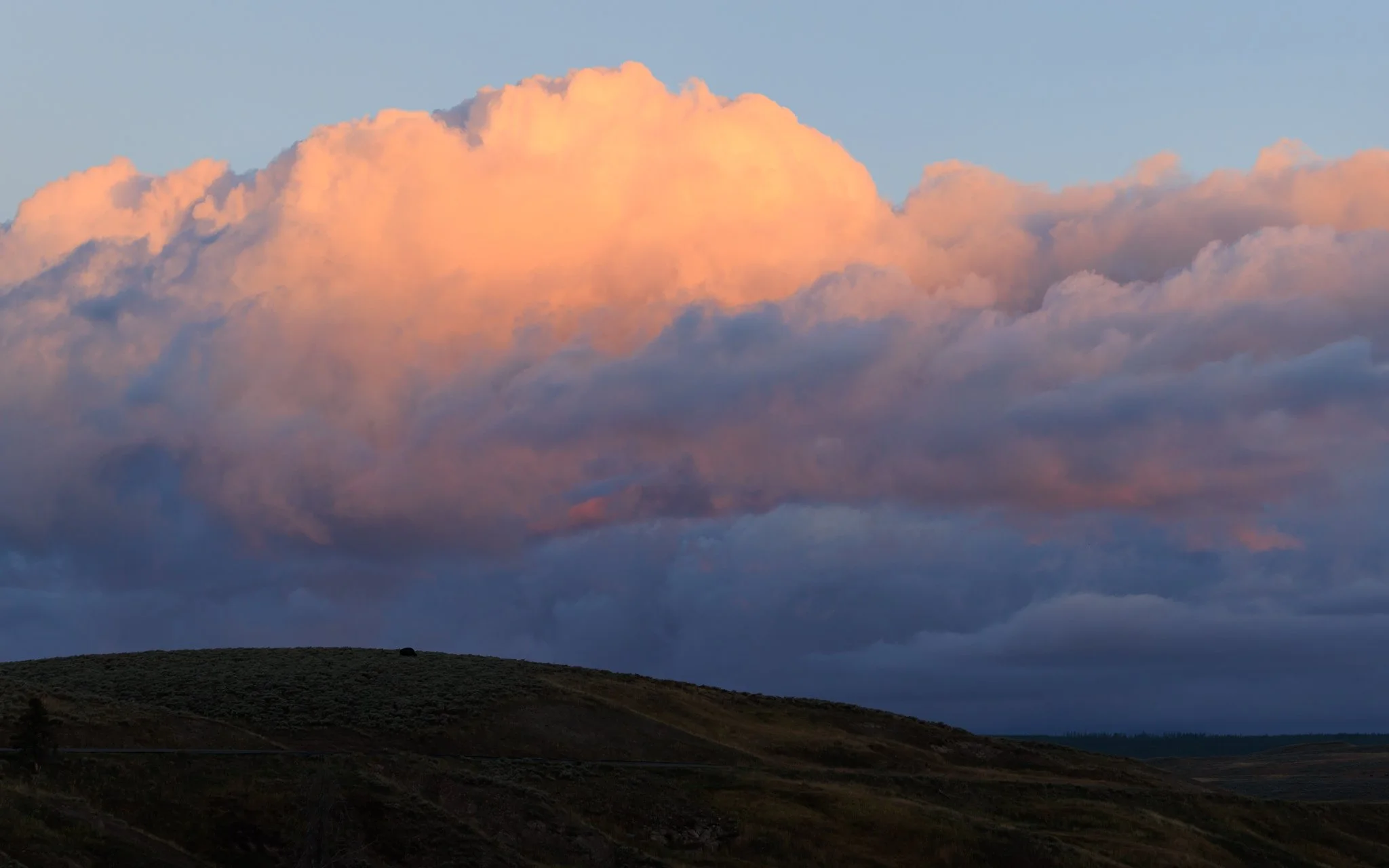 Towering clouds glowing peach and coral at dawn above a dark sagebrush hill in Hayden Valley, Yellowstone National Park.