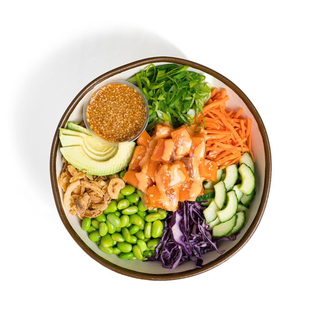 A bowl of poke with various vegetables, including avocado, edamame, shredded carrots, sliced cucumbers, purple cabbage, chopped green onions, and a small container of sesame seed dressing.