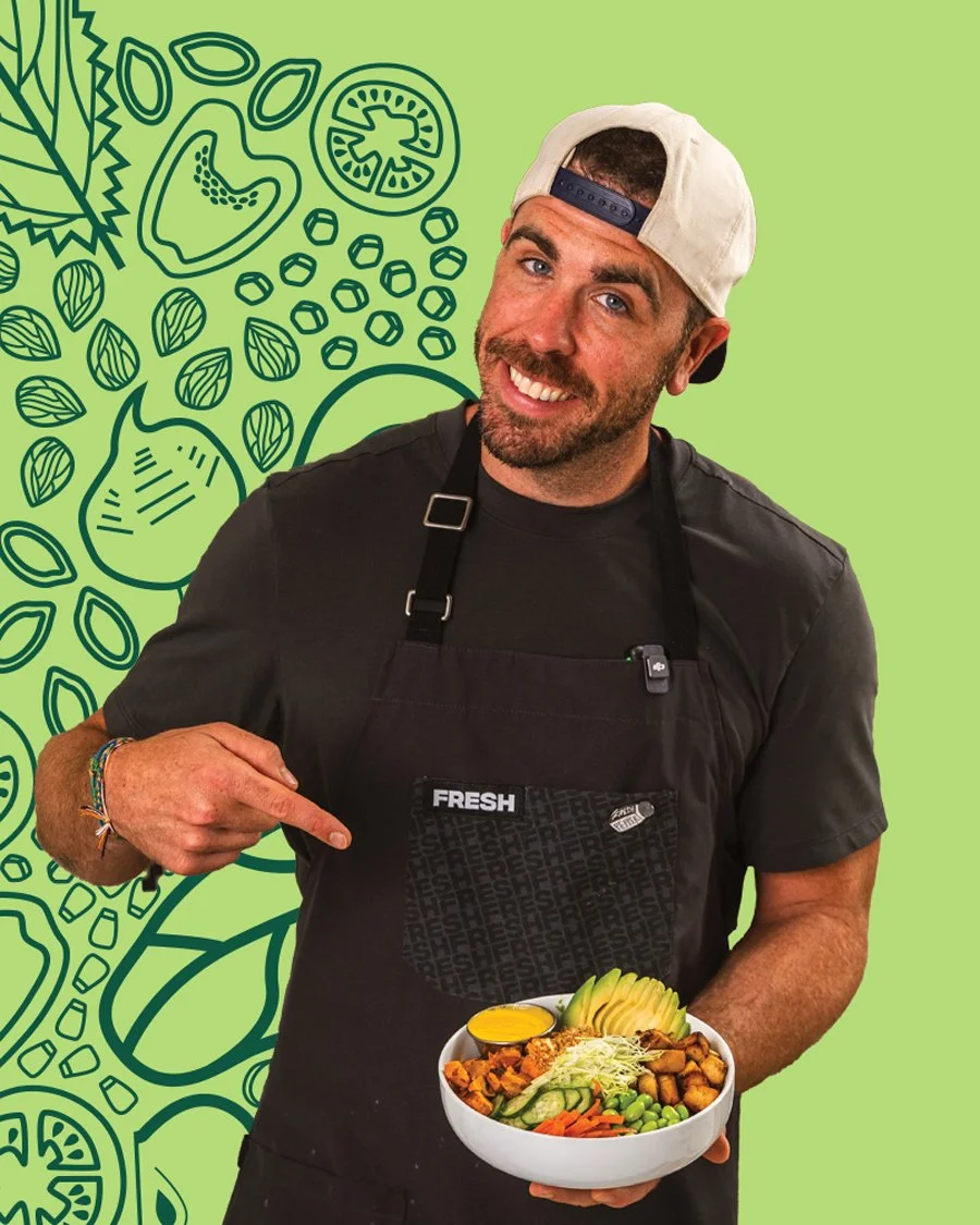 A smiling man wearing a black apron labeled 'FRESH' and a backwards white cap, holding a bowl of colorful salad with a lemon wedge, sliced avocado, and mixed vegetables, against a green background with black line drawings of various vegetables and herbs.