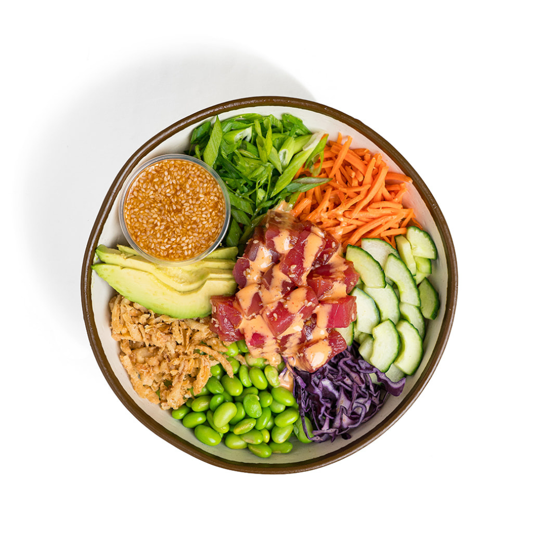 A colorful poke bowl with chopped vegetables, tuna, ginger, avocado, edamame, shredded purple cabbage, shredded carrots, sliced cucumbers, green onions, and a small bowl of sesame sauce.