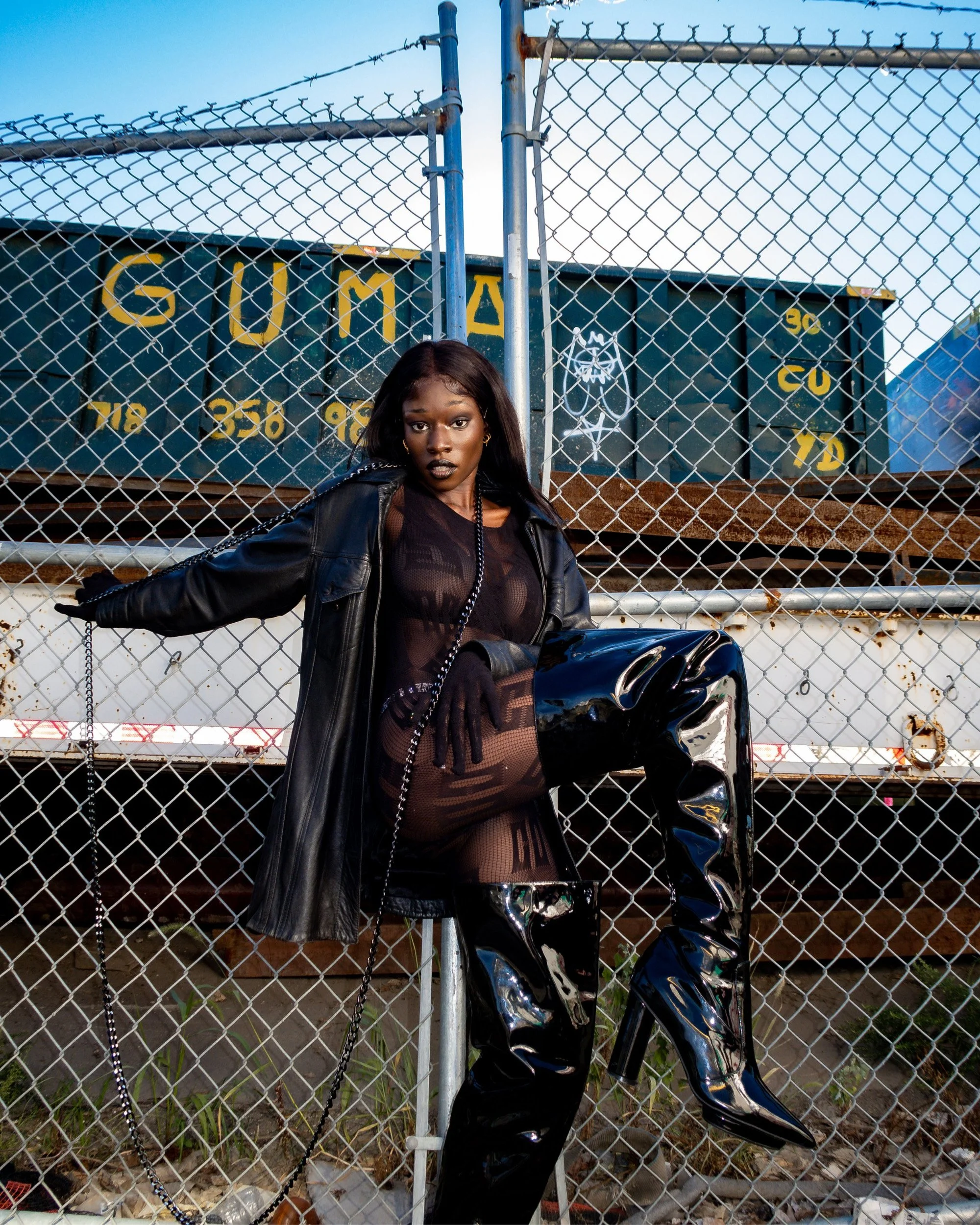 A woman with dark hair, wearing a sheer black outfit, black leather jacket, fishnet stockings, and shiny black boots, stands behind a chain-link fence with a green train car in the background. The train car has yellow and white graffiti and rust.