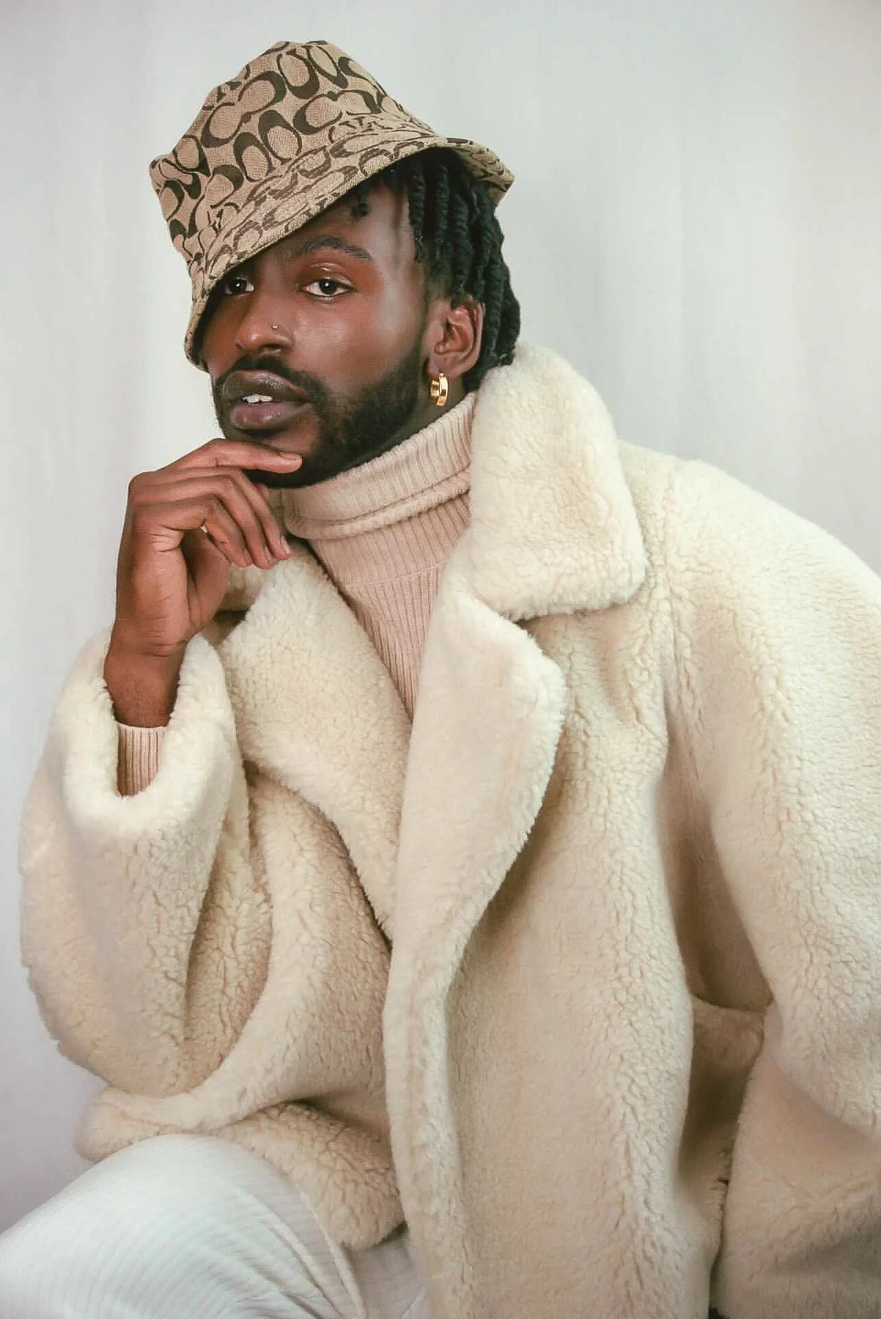 A young man with a beard and braided hair wearing a beige fuzzy coat, a tan turtleneck sweater, a patterned bucket hat, a single earring, and a nose ring, sitting against a plain light background.