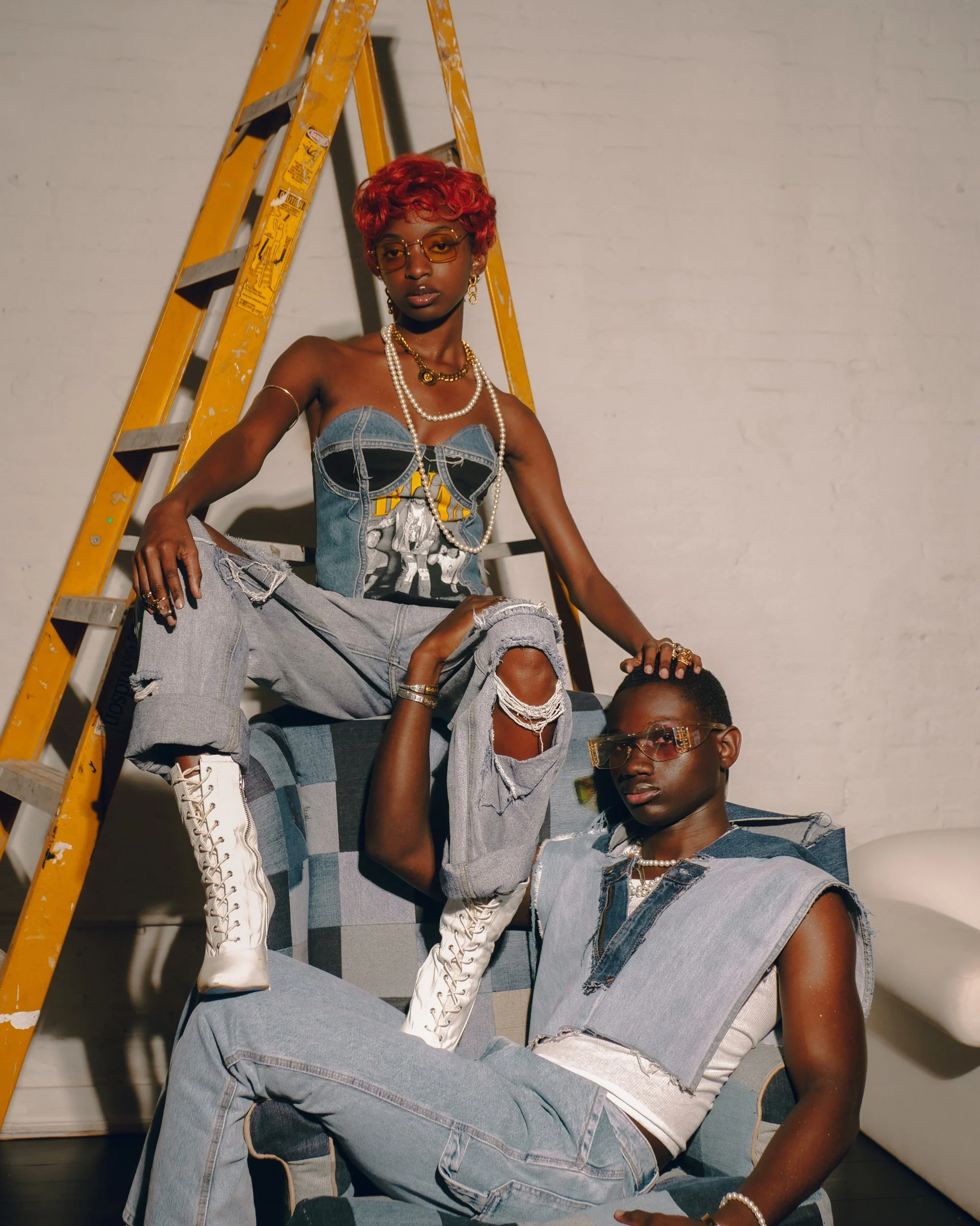 Four women posing on and around a yellow ladder against a white brick wall. They are dressed in denim outfits, wearing sunglasses, jewelry, and stylish accessories.