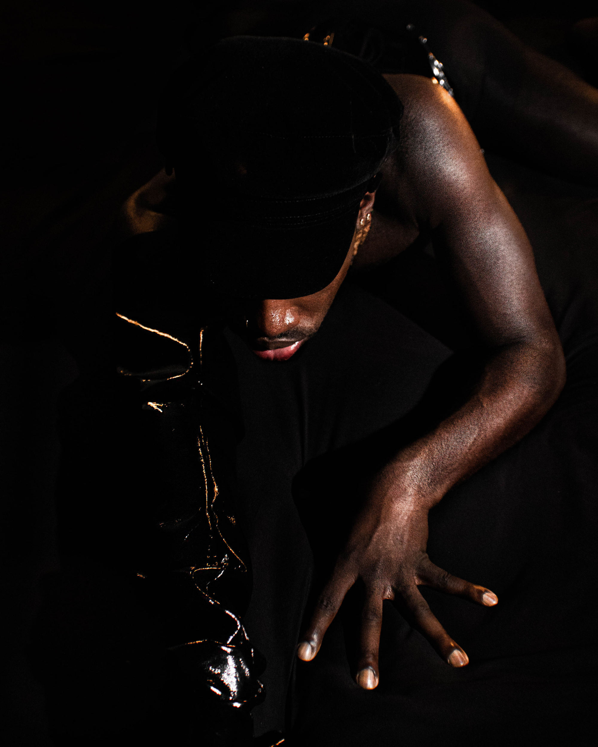 A person with dark skin wearing a black cap and a black top, lying on their stomach on a dark surface, with a silver chain accessory around their neck, and their face partly visible.