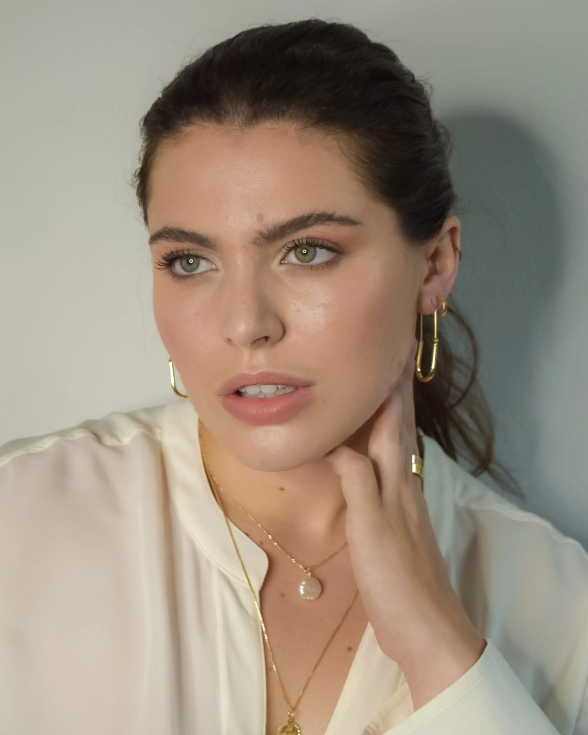 Close-up of a woman with dark hair, light green eyes, wearing gold hoop earrings, a pearl necklace, and a cream-colored top, touching her face with her right hand, against a plain, light background.