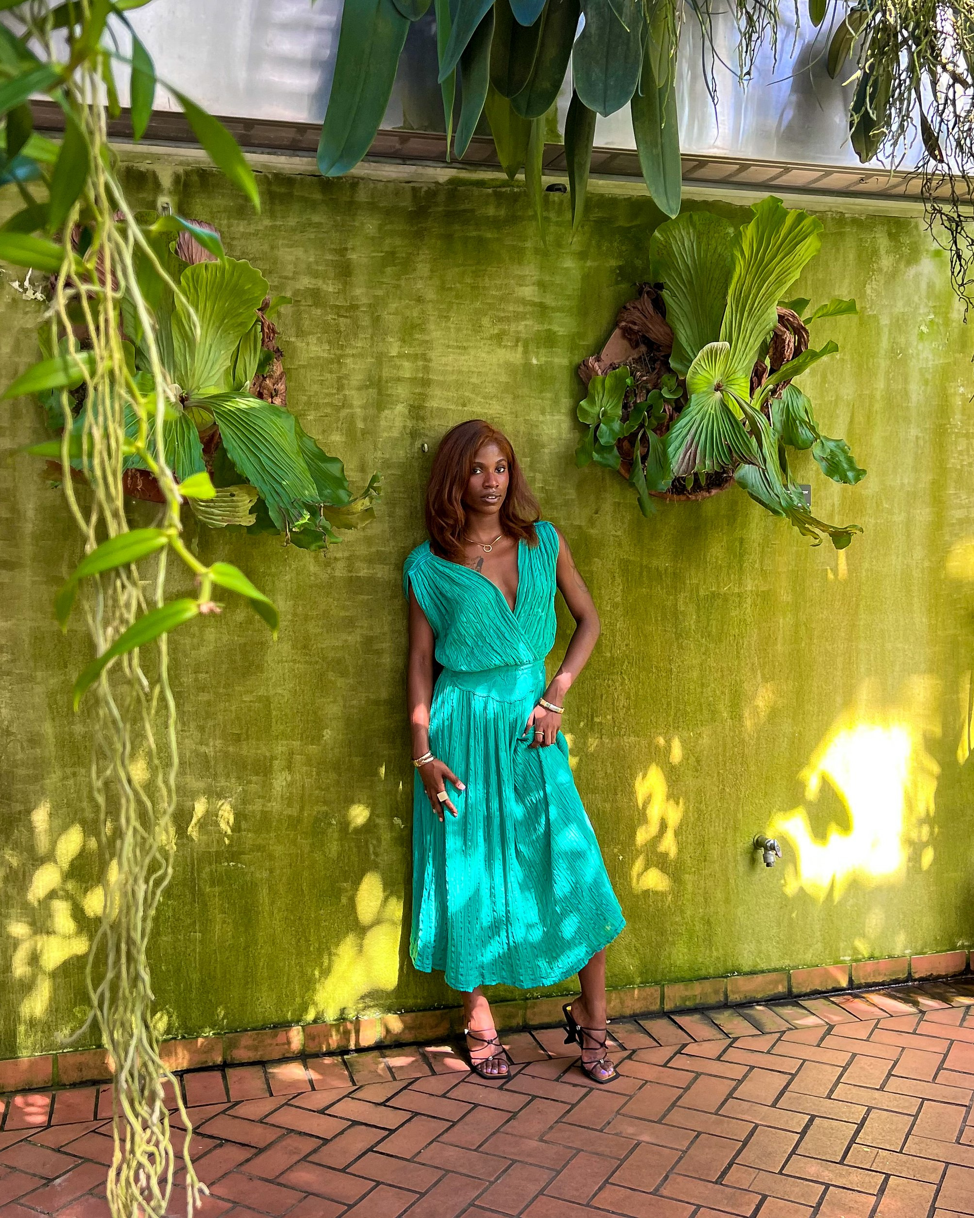 A woman in a turquoise dress with black high heels standing in front of a green textured wall with large tropical plants hanging above. She has reddish-brown hair and jewelry on her wrists and neck.