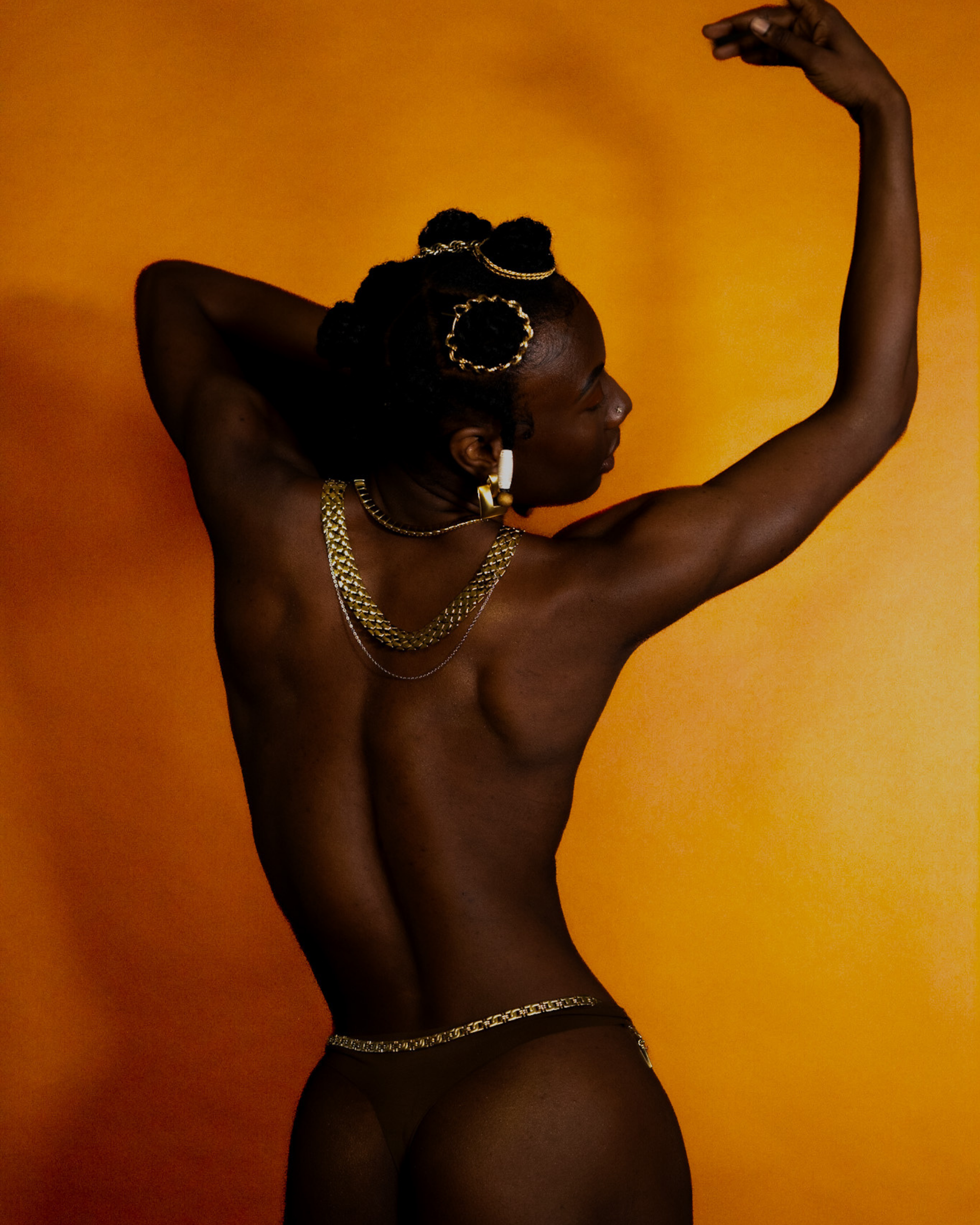 A photograph of a dark-skinned woman with braided hair, adorned with gold jewelry, striking a pose against a vibrant orange background.