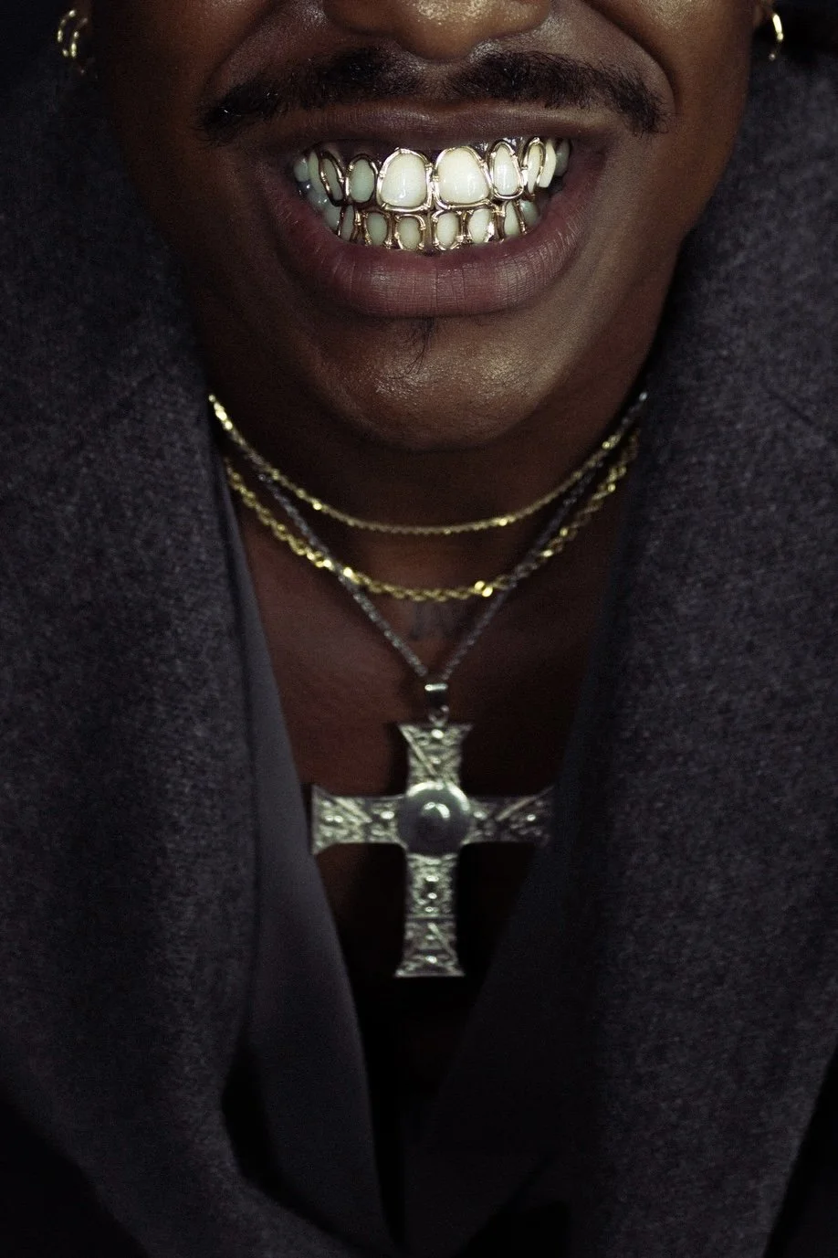 Close-up of a smiling person showing gold and diamond grilles over their teeth, wearing layered gold necklaces and a large silver cross pendant.