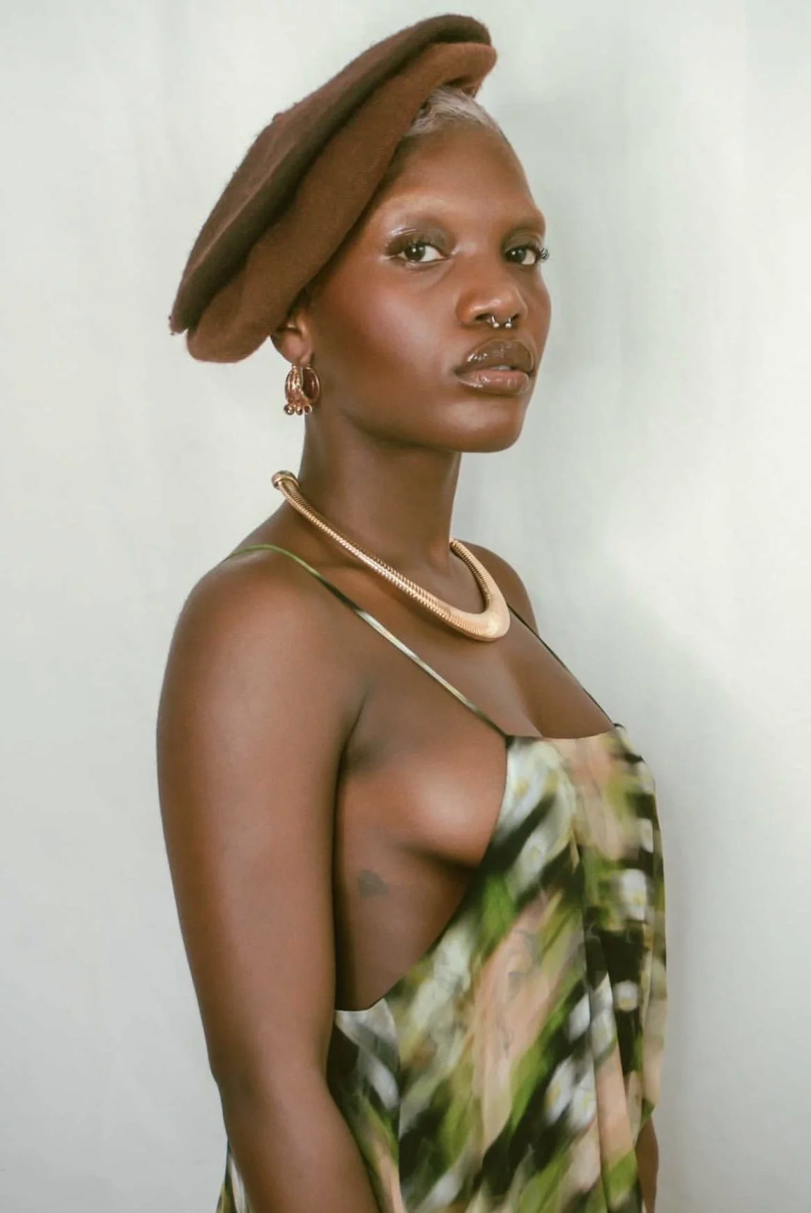 A woman with dark skin and silver hair styled in a bun, wearing a brown beret, gold jewelry, and a patterned dress with spaghetti straps, standing against a plain light-colored background.